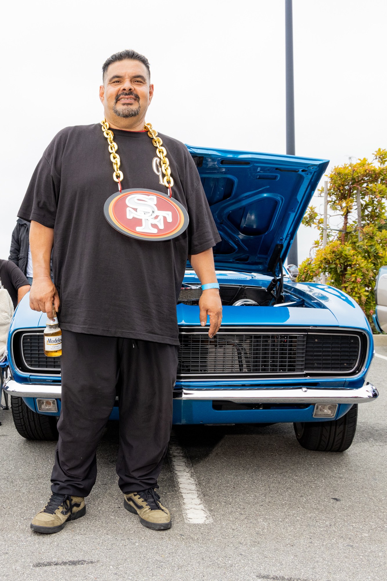 A man stands in front of a blue car with its hood open, wearing a large chain with a football team logo. Other people relax in chairs around a red convertible and another vehicle.