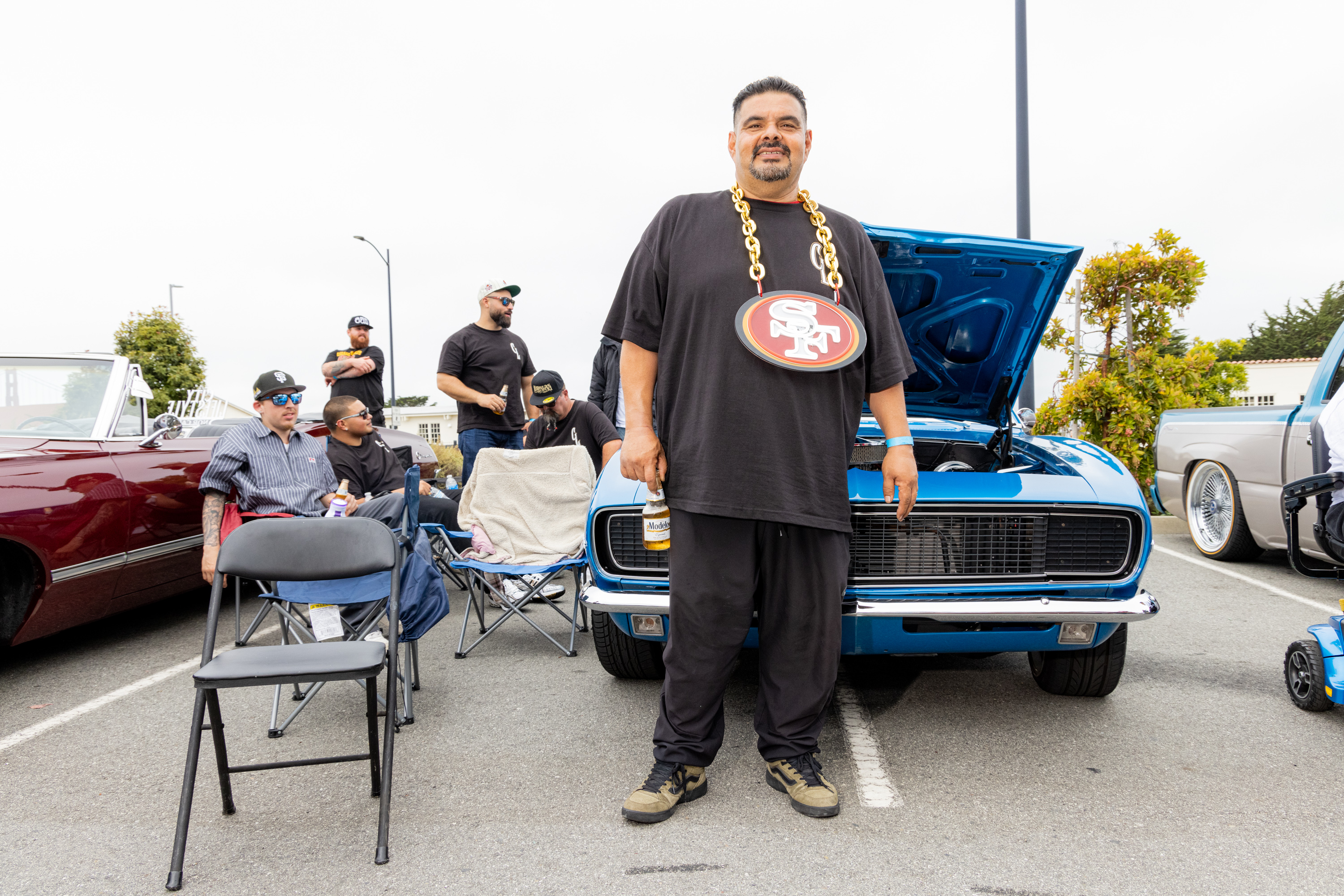 A man stands in front of a blue car with its hood open, wearing a large chain with a football team logo. Other people relax in chairs around a red convertible and another vehicle.