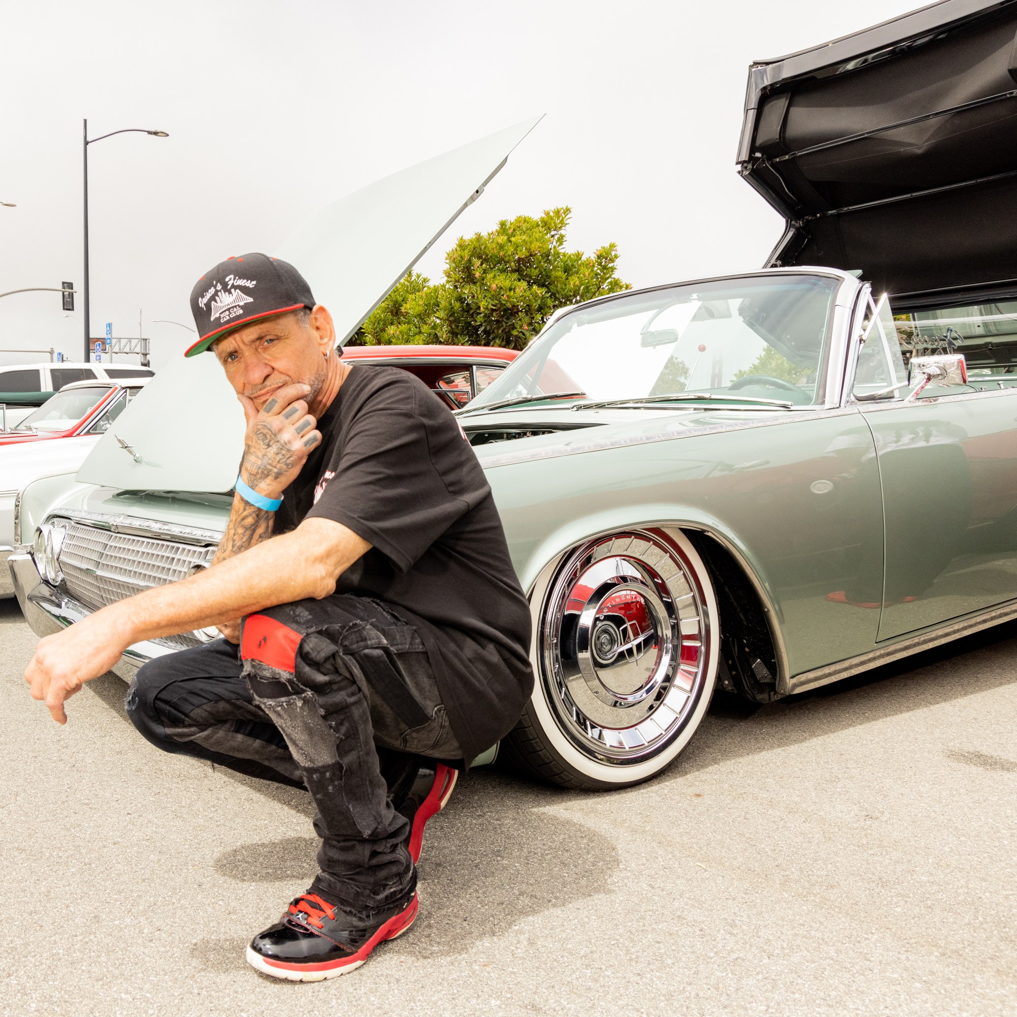 A man squats near vintage cars with hoods open. He wears a black cap, shirt, and red-accented shoes. Green folding chairs and people are in the background.