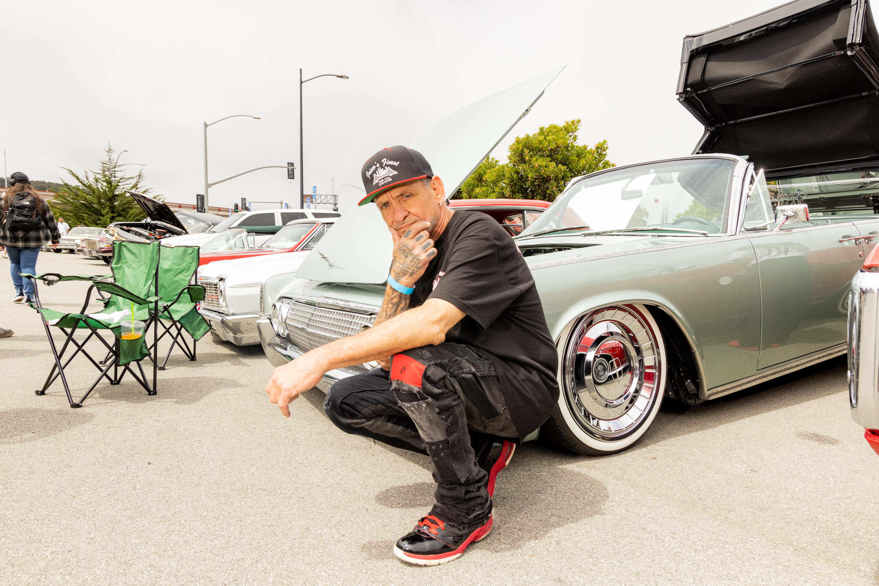 A man squats near vintage cars with hoods open. He wears a black cap, shirt, and red-accented shoes. Green folding chairs and people are in the background.