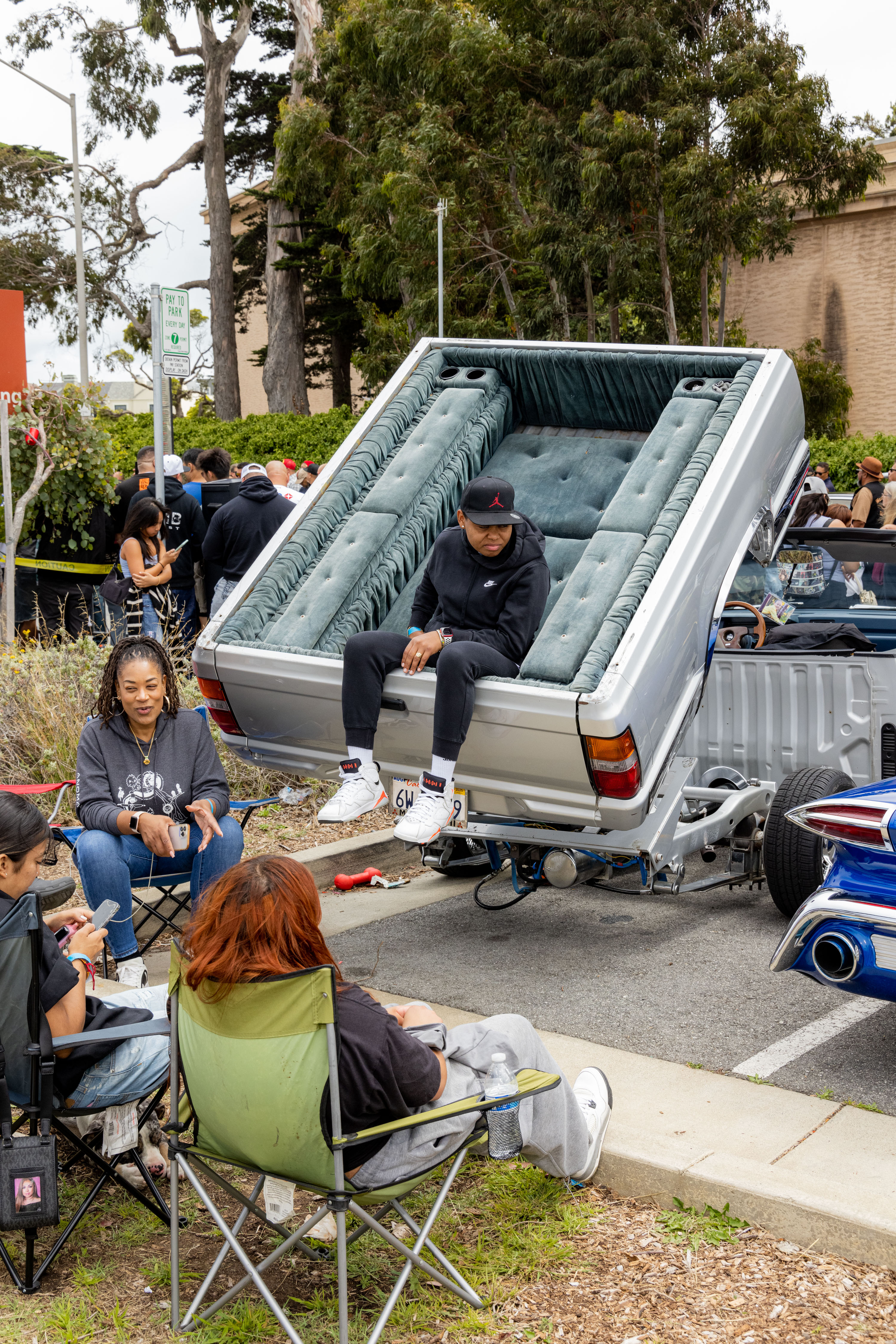 A man sits on a modified car with a flipped vertical bed, surrounded by three other people sitting in lawn chairs on the sidewalk. Trees and a street sign are visible.