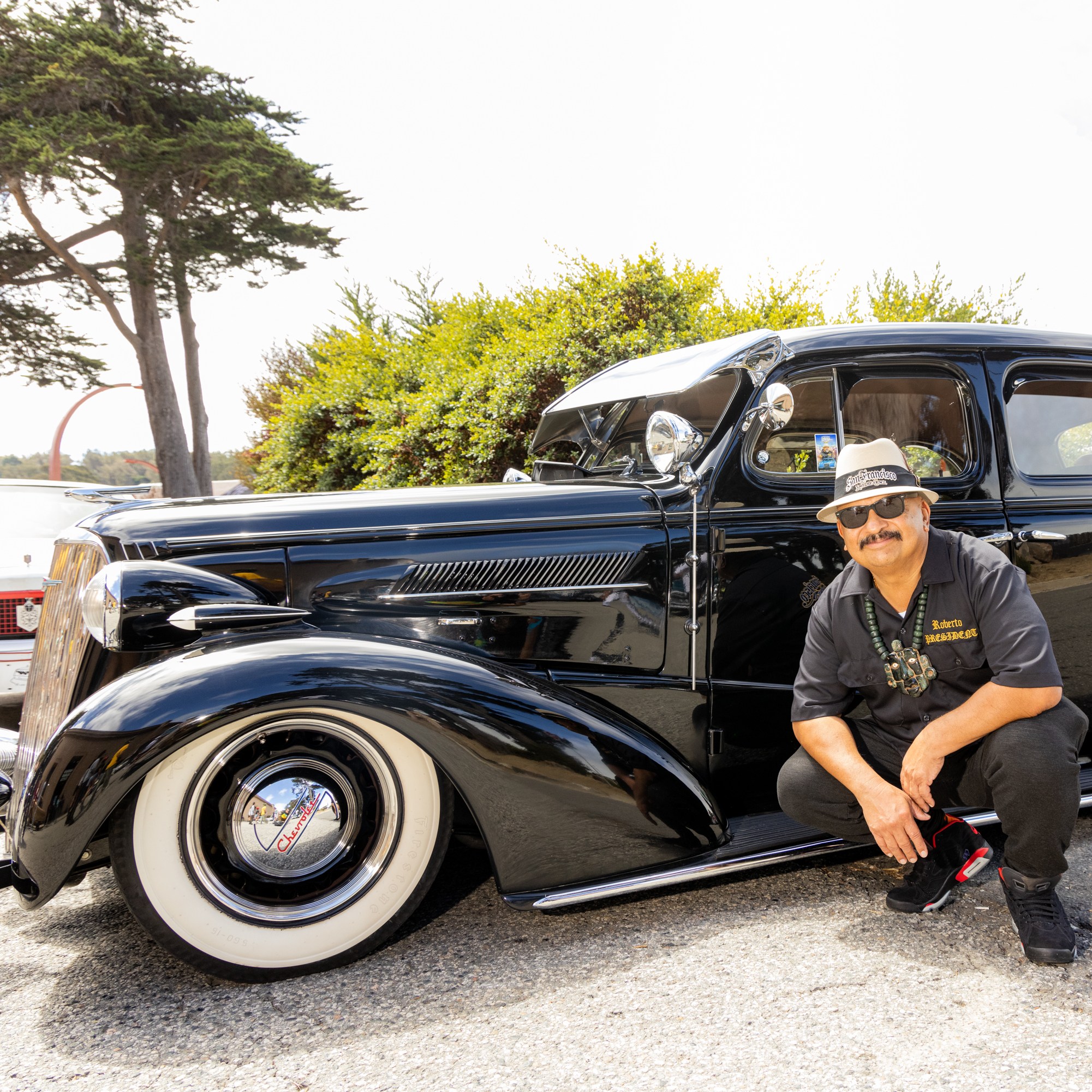 A man kneels beside a polished black vintage car with whitewall tires. He wears a hat, sunglasses, and a black shirt. Trees and other classic cars are in the background.