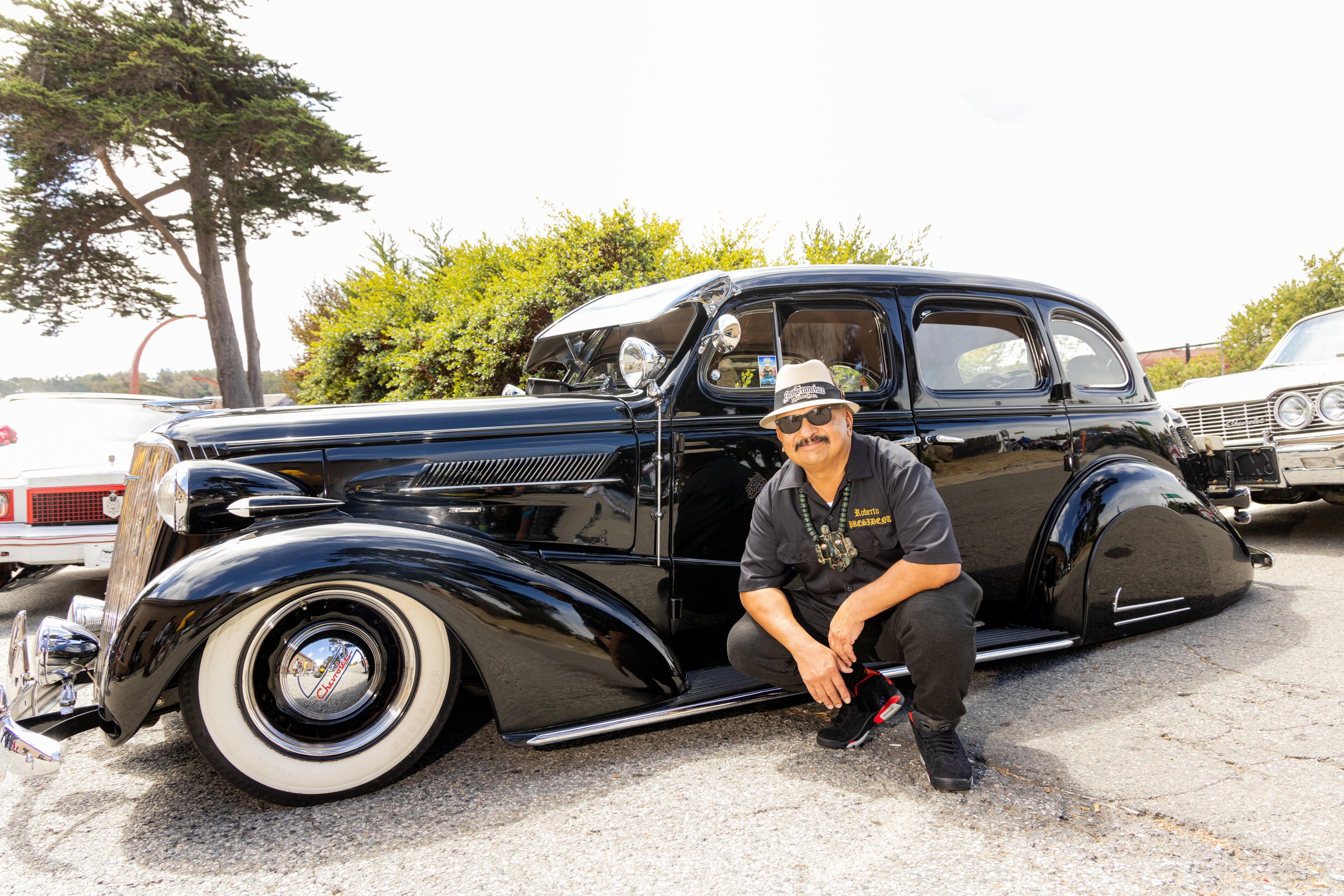 A man kneels beside a polished black vintage car with whitewall tires. He wears a hat, sunglasses, and a black shirt. Trees and other classic cars are in the background.