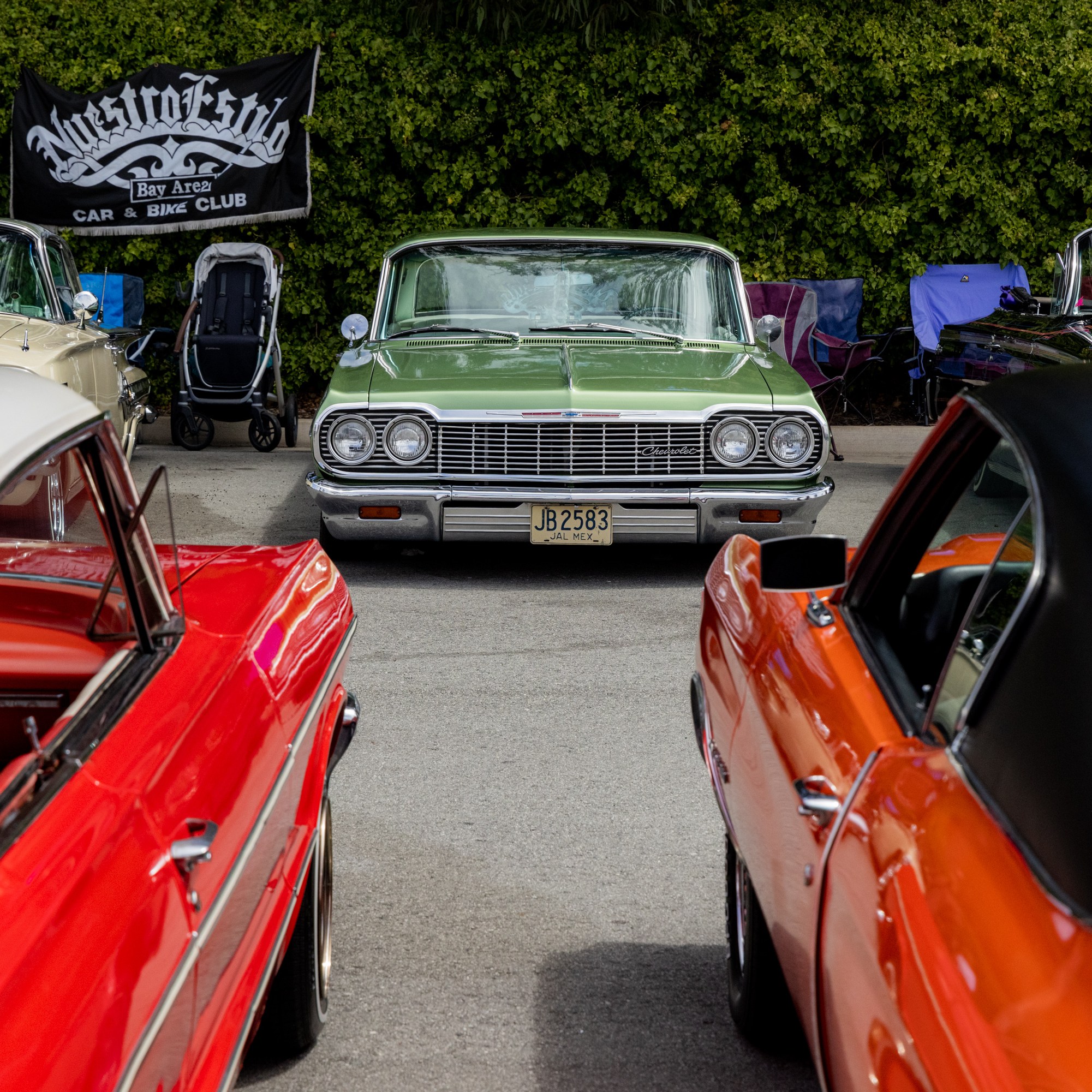 A green vintage car is parked facing forward at a car show, surrounded by other colorful classic cars, with people and a &quot;Maestro Klub&quot; banner in the background.