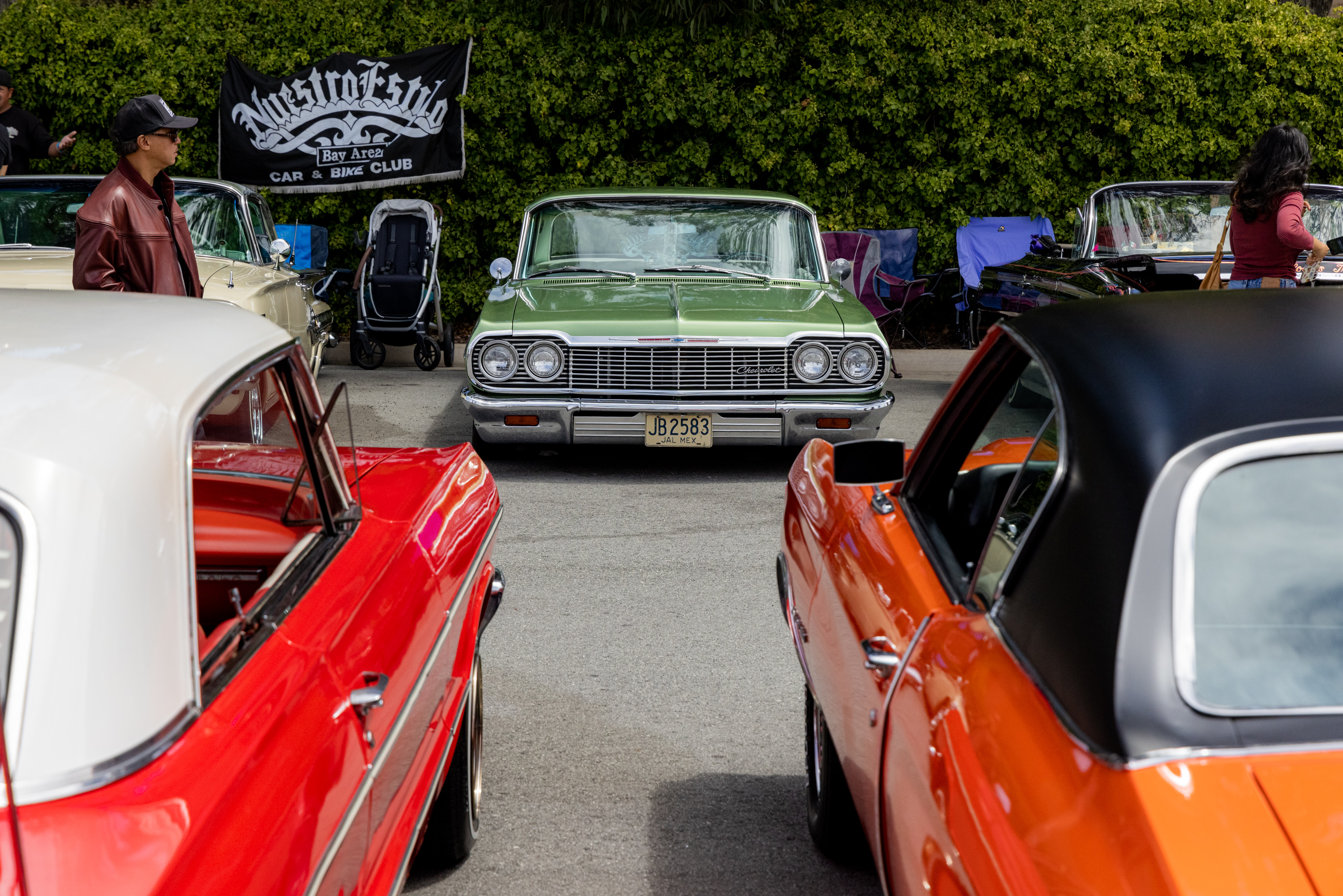 A green vintage car is parked facing forward at a car show, surrounded by other colorful classic cars, with people and a &quot;Maestro Klub&quot; banner in the background.