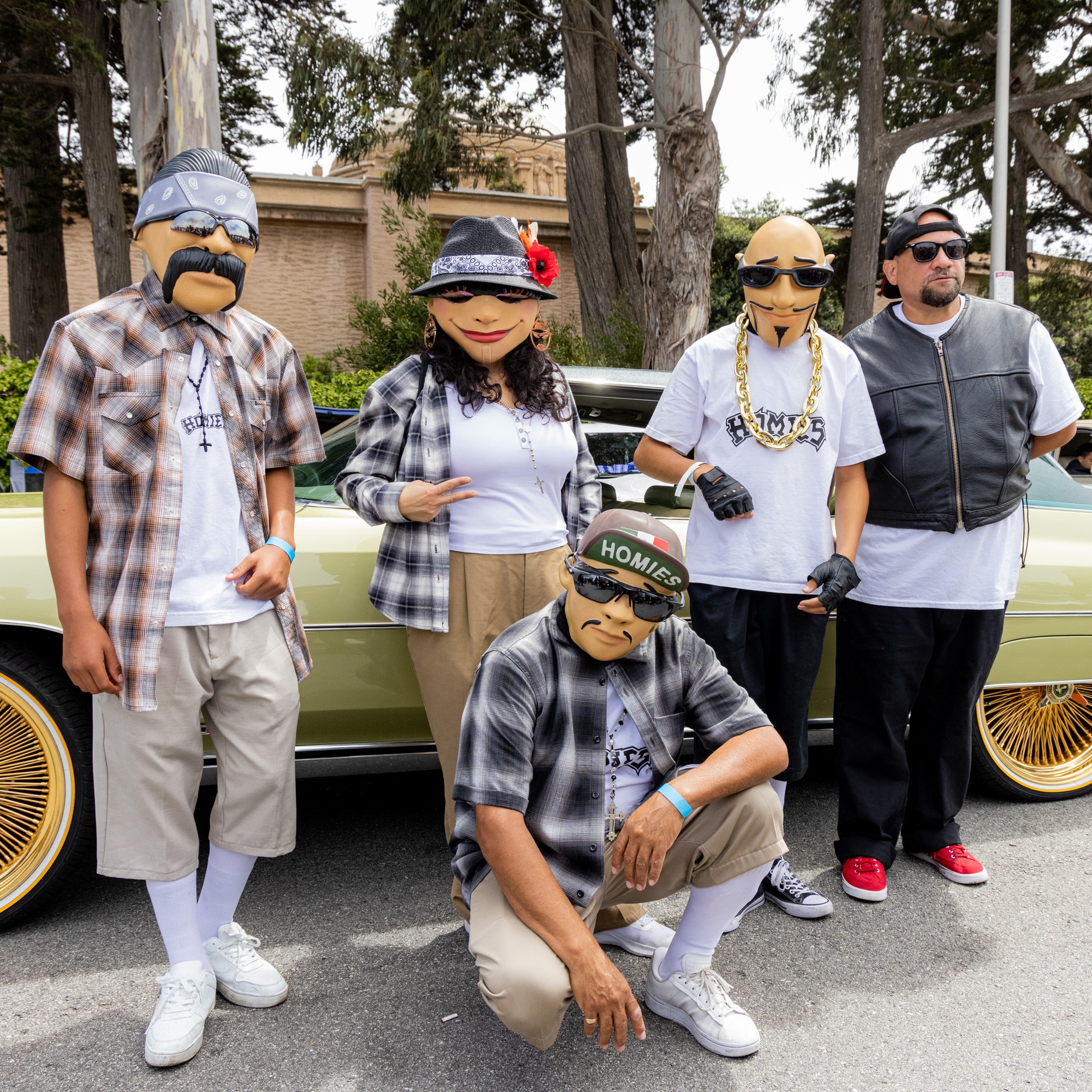 Five people wearing masks and casual clothing pose in front of a green vintage car with gold rims, outdoors on a street lined with trees, and spectators in the background.