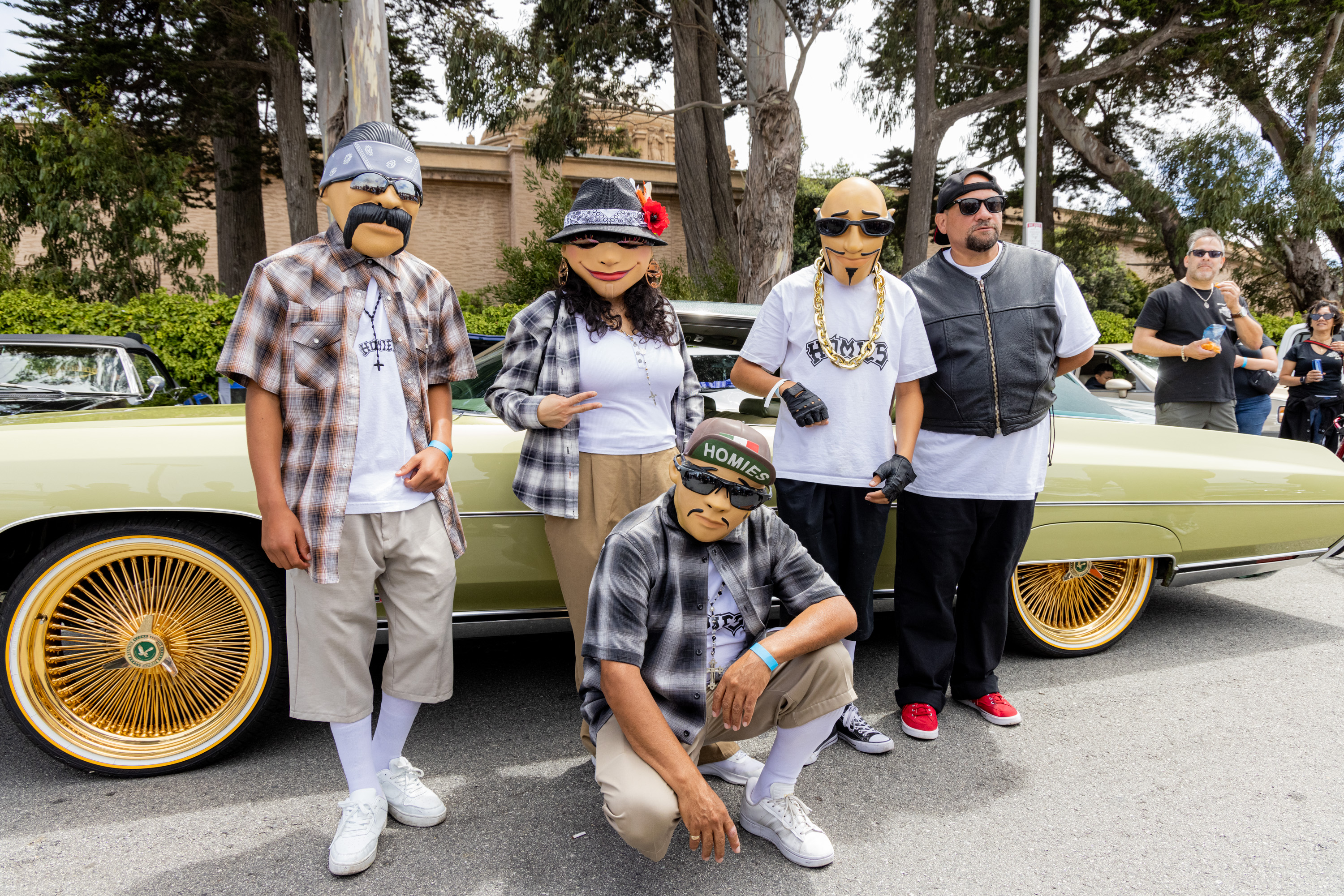 Five people wearing masks and casual clothing pose in front of a green vintage car with gold rims, outdoors on a street lined with trees, and spectators in the background.