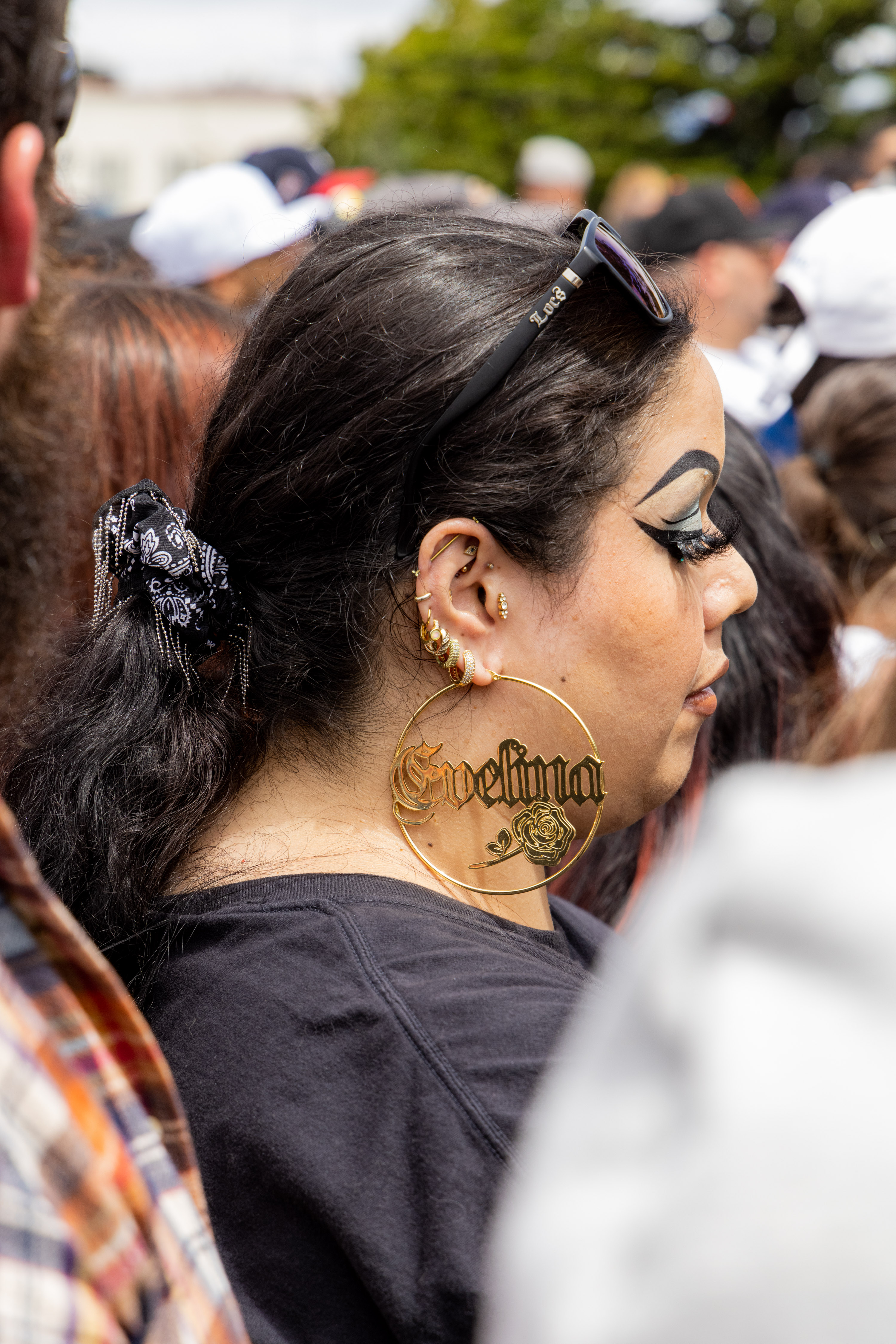 A woman in a crowd has long dark hair tied with a black bandanna, large gold hoop earrings with &quot;Evelina&quot; and a rose, multiple ear piercings, bold makeup, and sunglasses on her head.