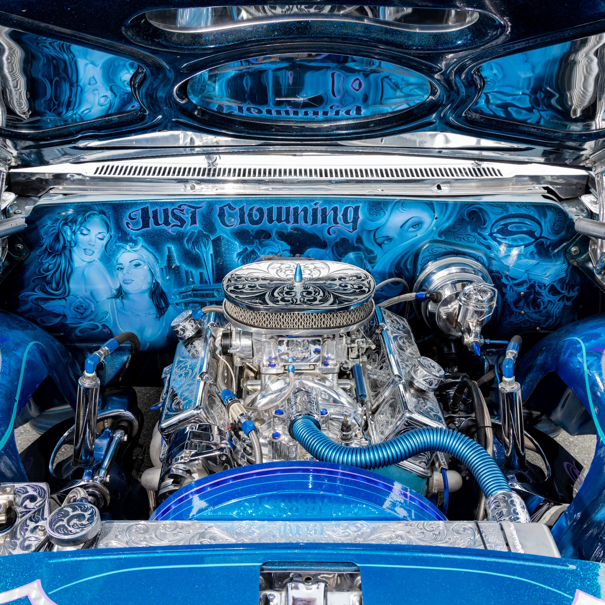 The image shows a customized car engine with detailed silver and blue components, artwork of women, and the phrase &quot;Just Clowning&quot; displayed inside the hood.