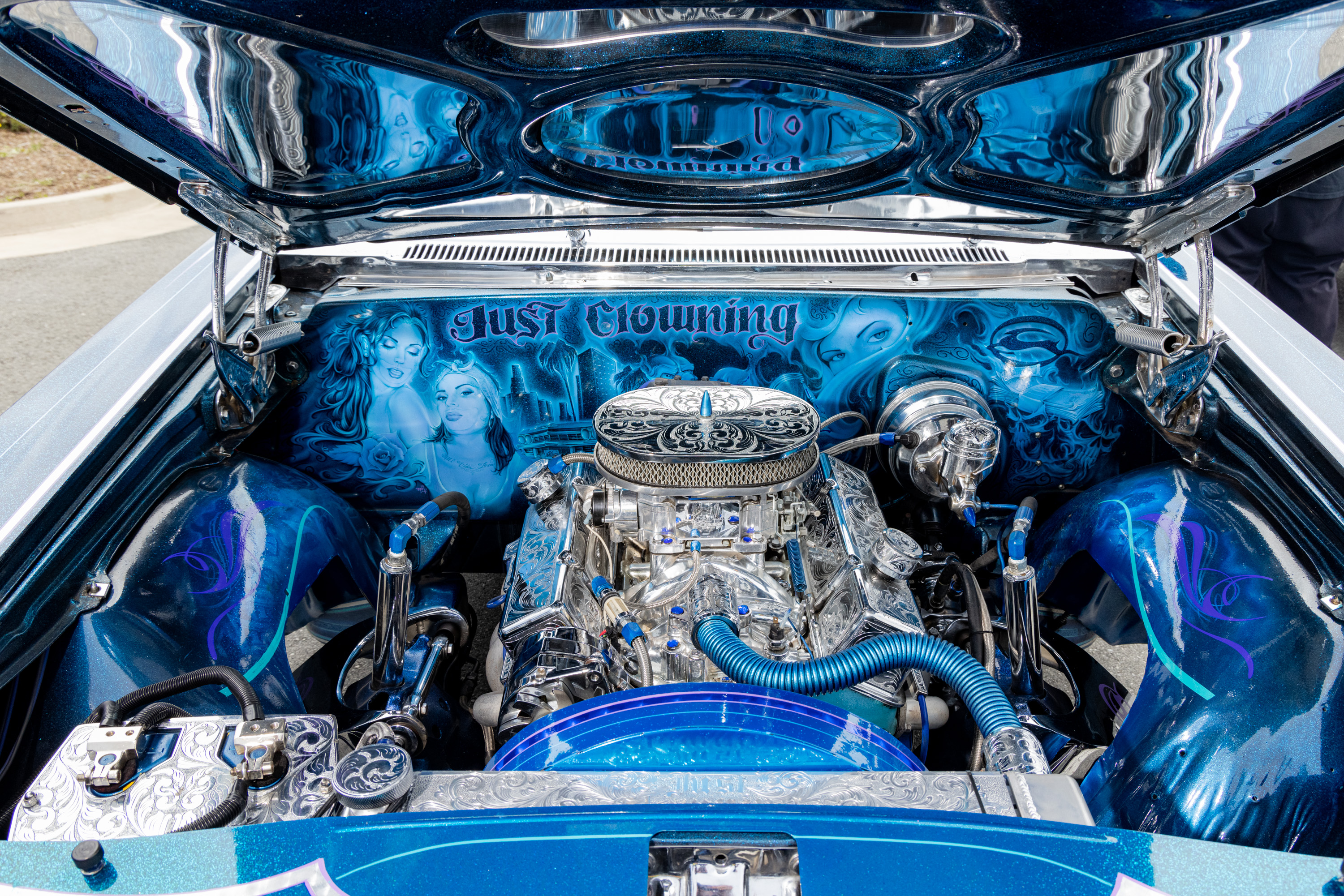 The image shows a customized car engine with detailed silver and blue components, artwork of women, and the phrase &quot;Just Clowning&quot; displayed inside the hood.