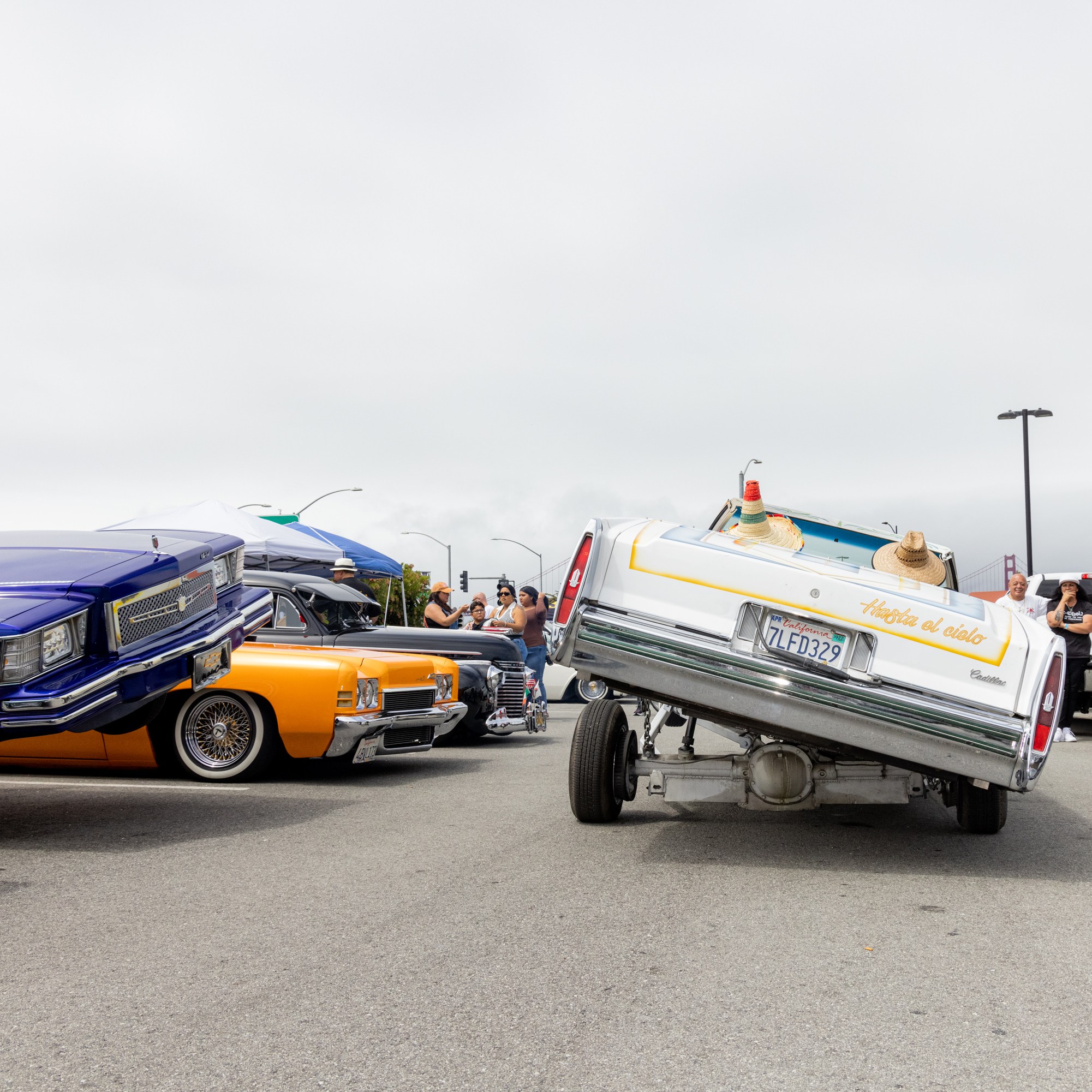 The image shows a white car performing a dramatic hydraulic lift stunt, tilted on one side. Nearby, colorful lowrider cars are parked, and people are watching and taking photos.