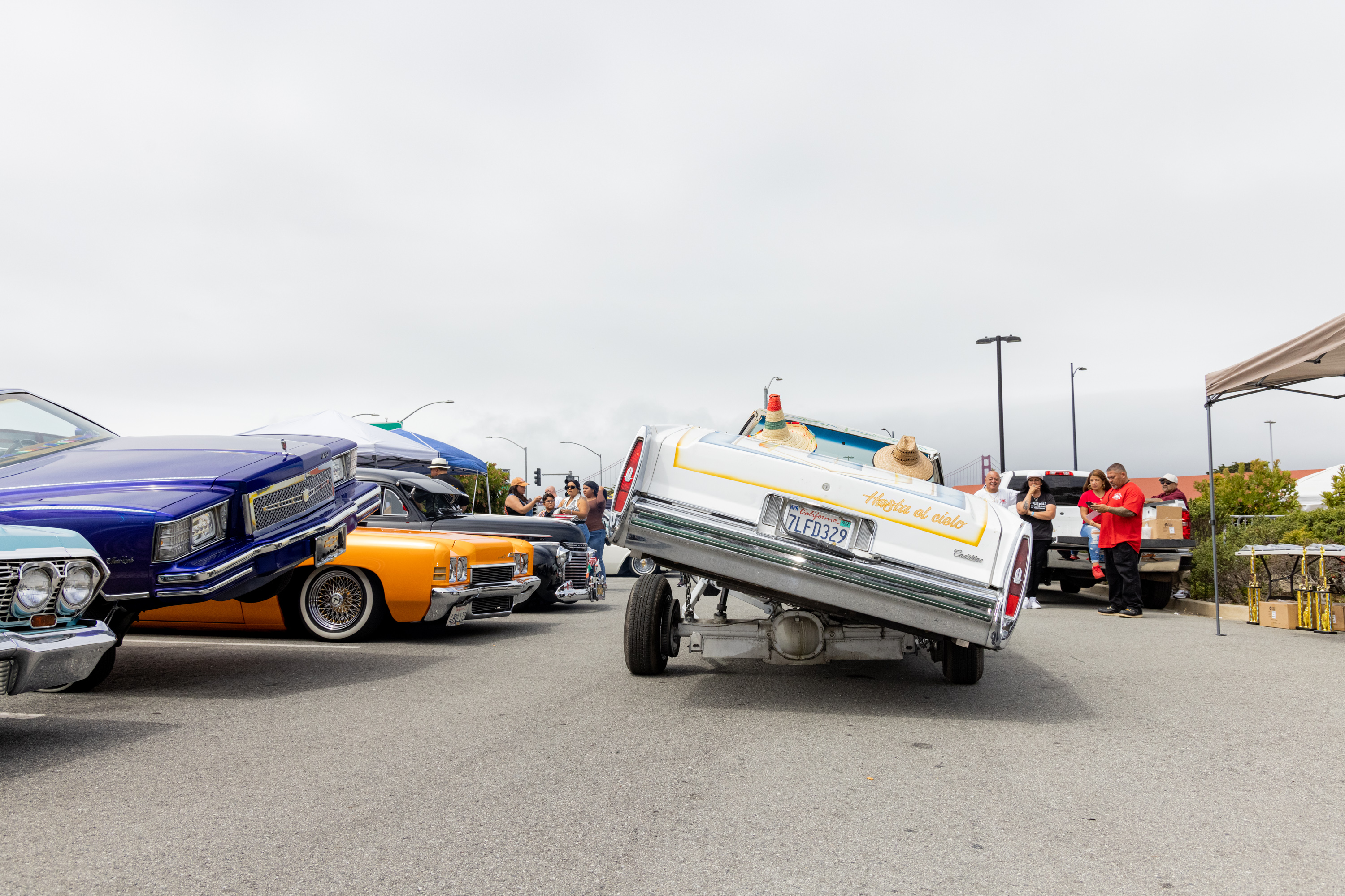 The image shows a white car performing a dramatic hydraulic lift stunt, tilted on one side. Nearby, colorful lowrider cars are parked, and people are watching and taking photos.