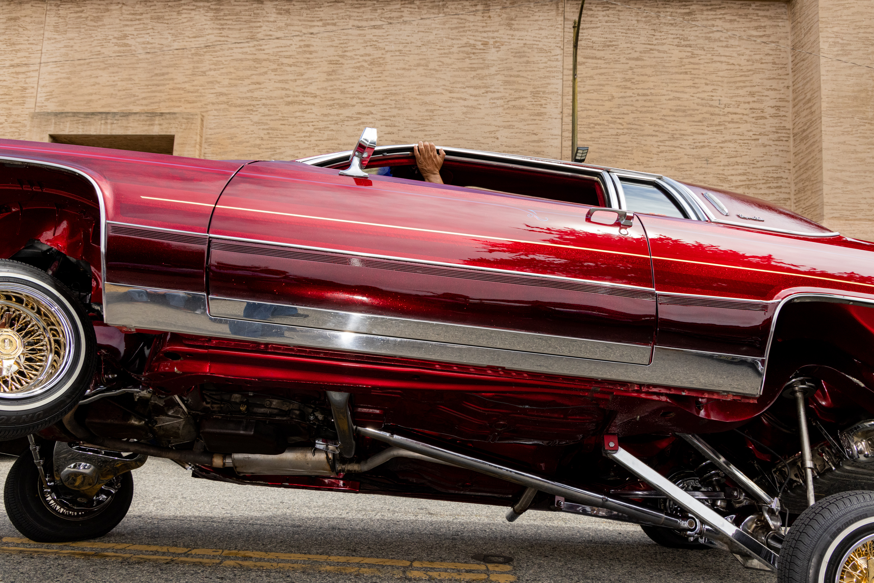 A red lowrider car with shiny chrome details is tilted diagonally, showing its undercarriage. A hand is visible from the rolled-down window, gripping it.