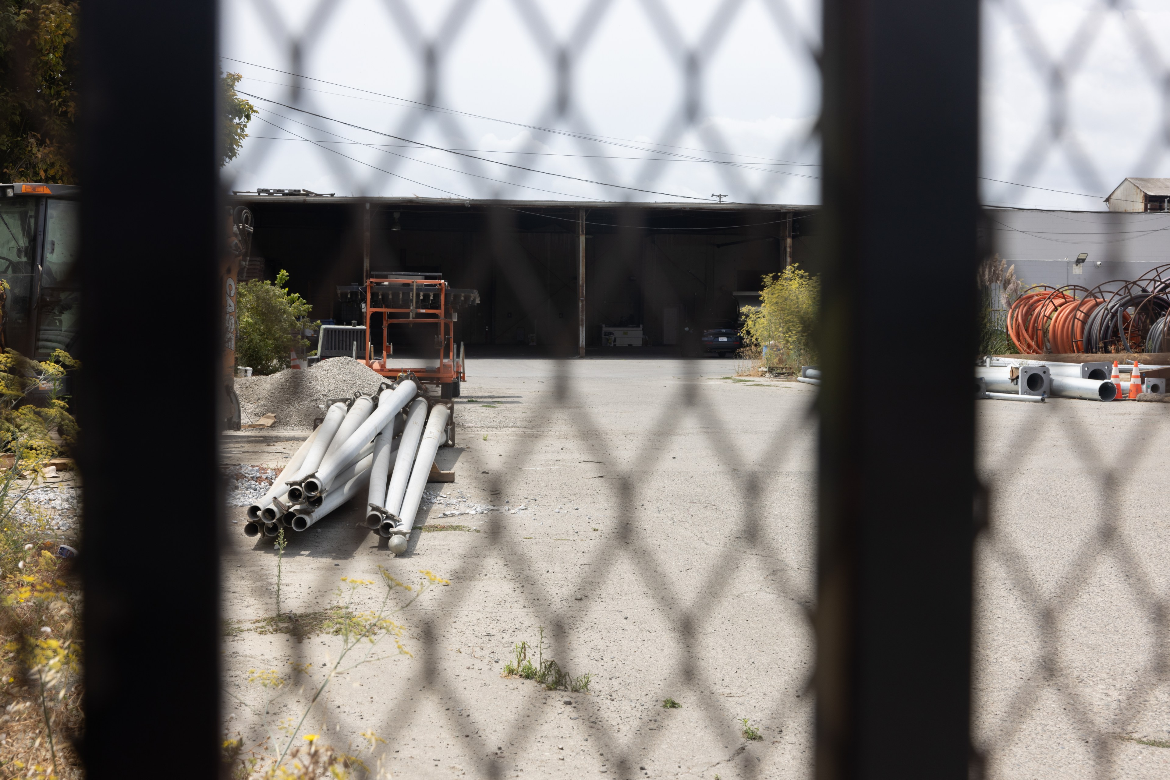 The image shows a fenced-off industrial yard with metal pipes, construction debris, and coiled cables. The scene is framed by a chain-link fence in the foreground.