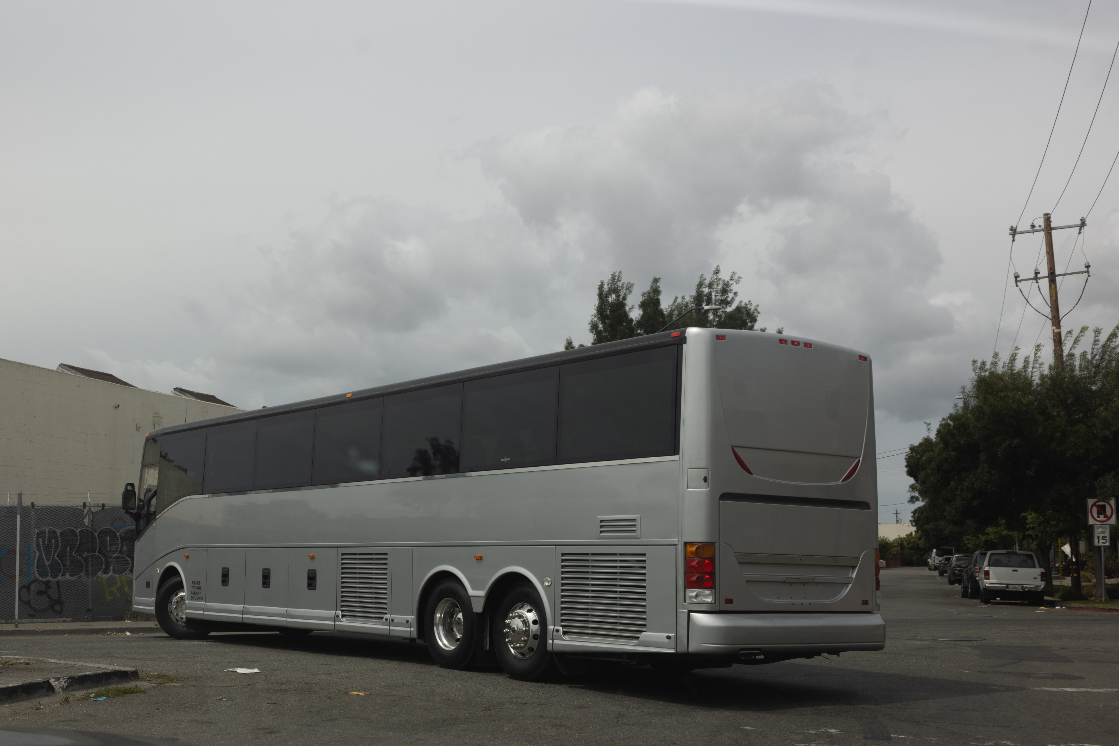 A large, silver tour bus is parked on a street. The sky is cloudy and there are trees, utility poles, and a building with graffiti in the background.