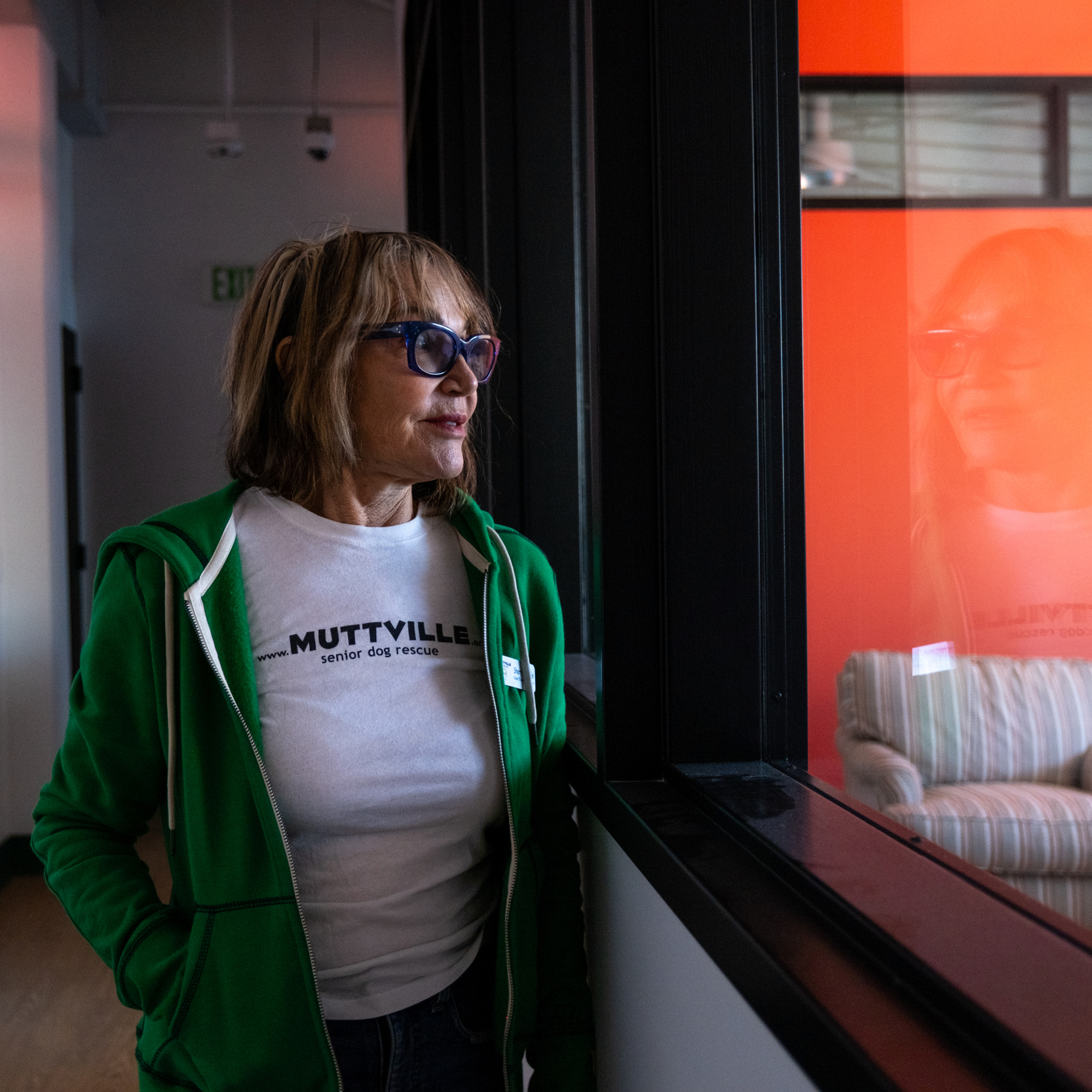 A woman in a green hoodie, white &quot;MUTTVILLE senior dog rescue&quot; t-shirt, and blue glasses stands by a window with a reflection. A striped chair is visible inside.