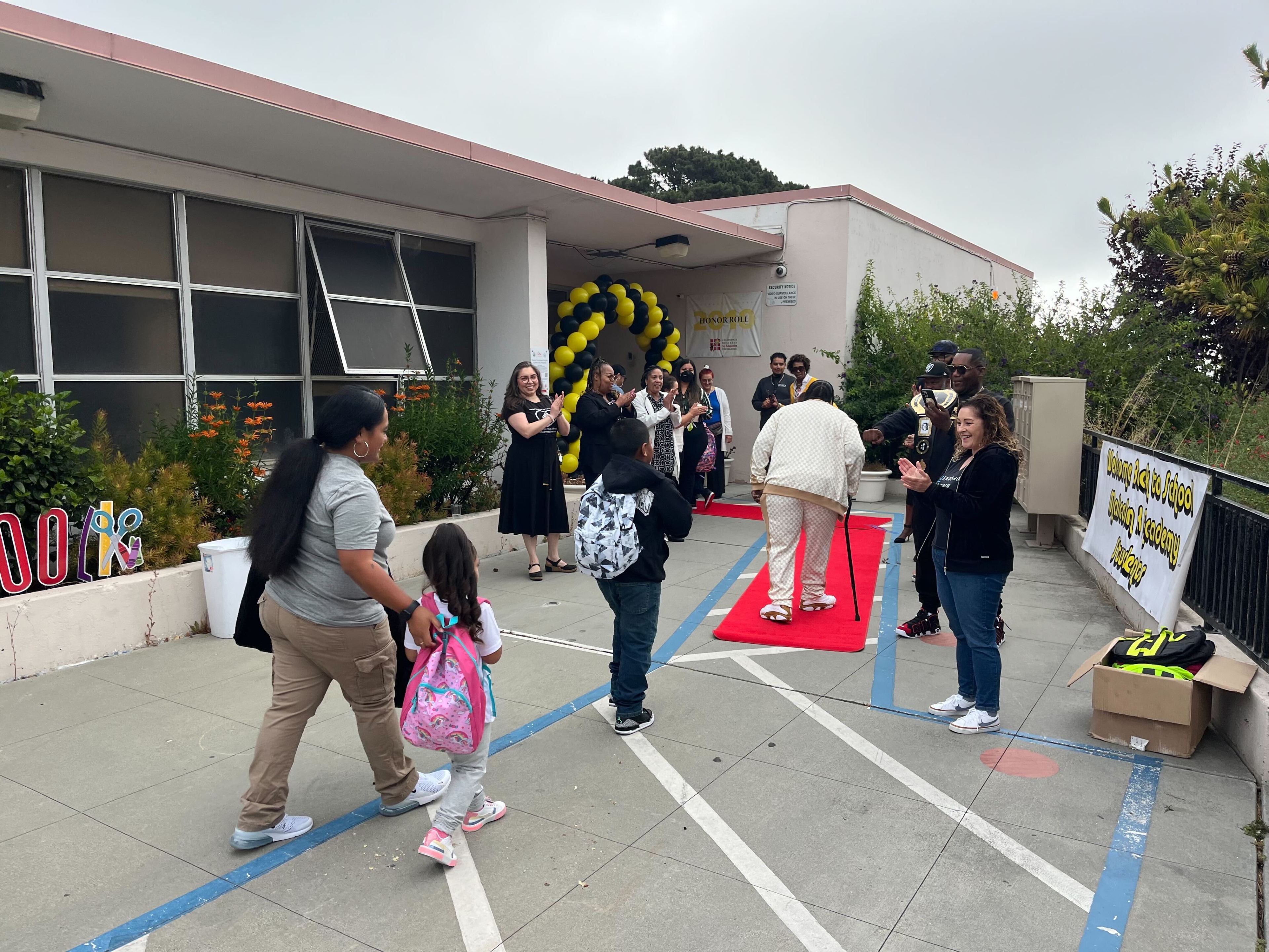 People gather outside a building with red carpet and balloon arch, greeting and applauding a person in white. A child in a pink backpack walks with an adult.