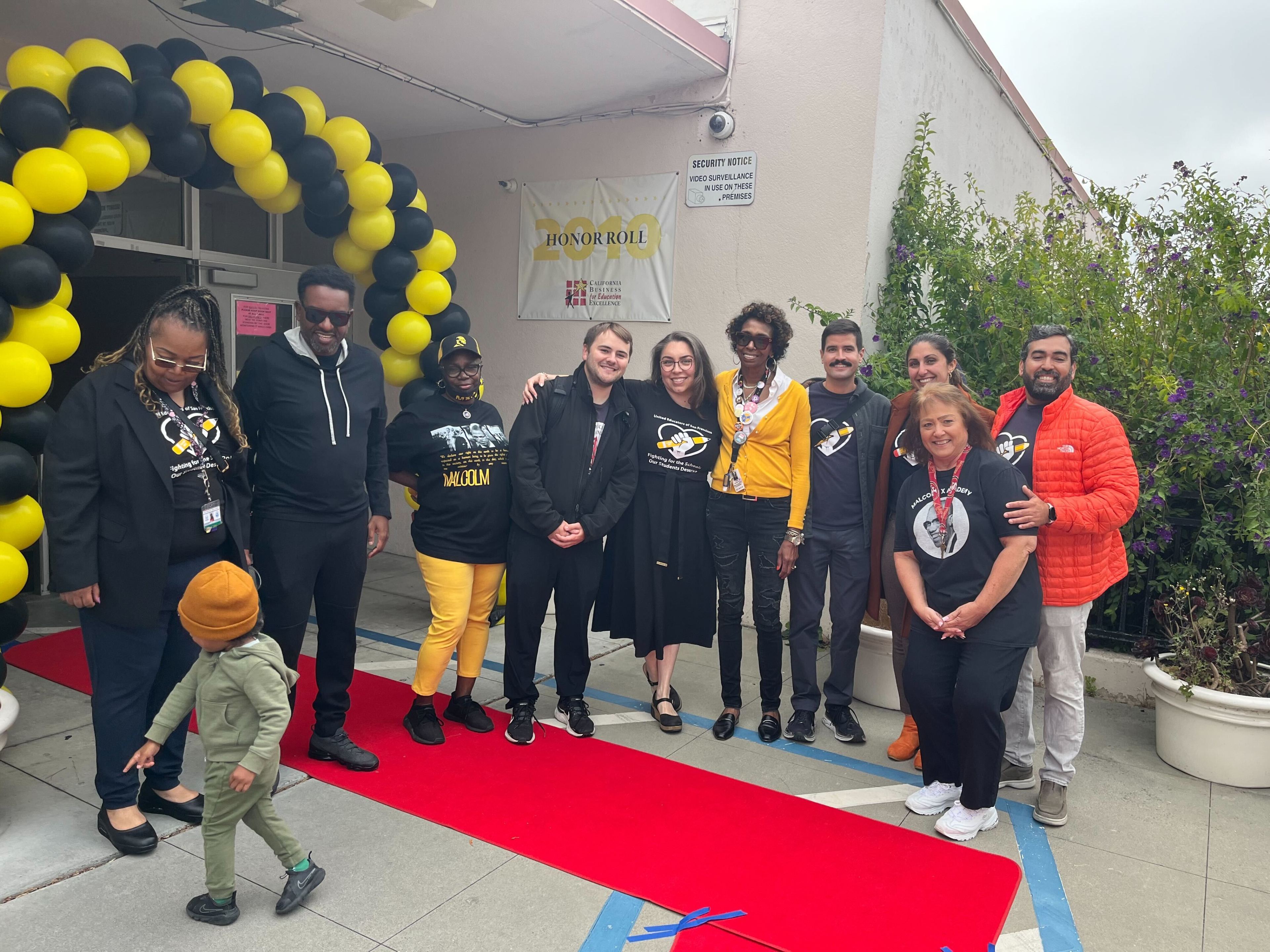 A group of eleven people are smiling and posing on a red carpet under a black and yellow balloon arch, with an "Honor Roll" sign and greenery in the background.