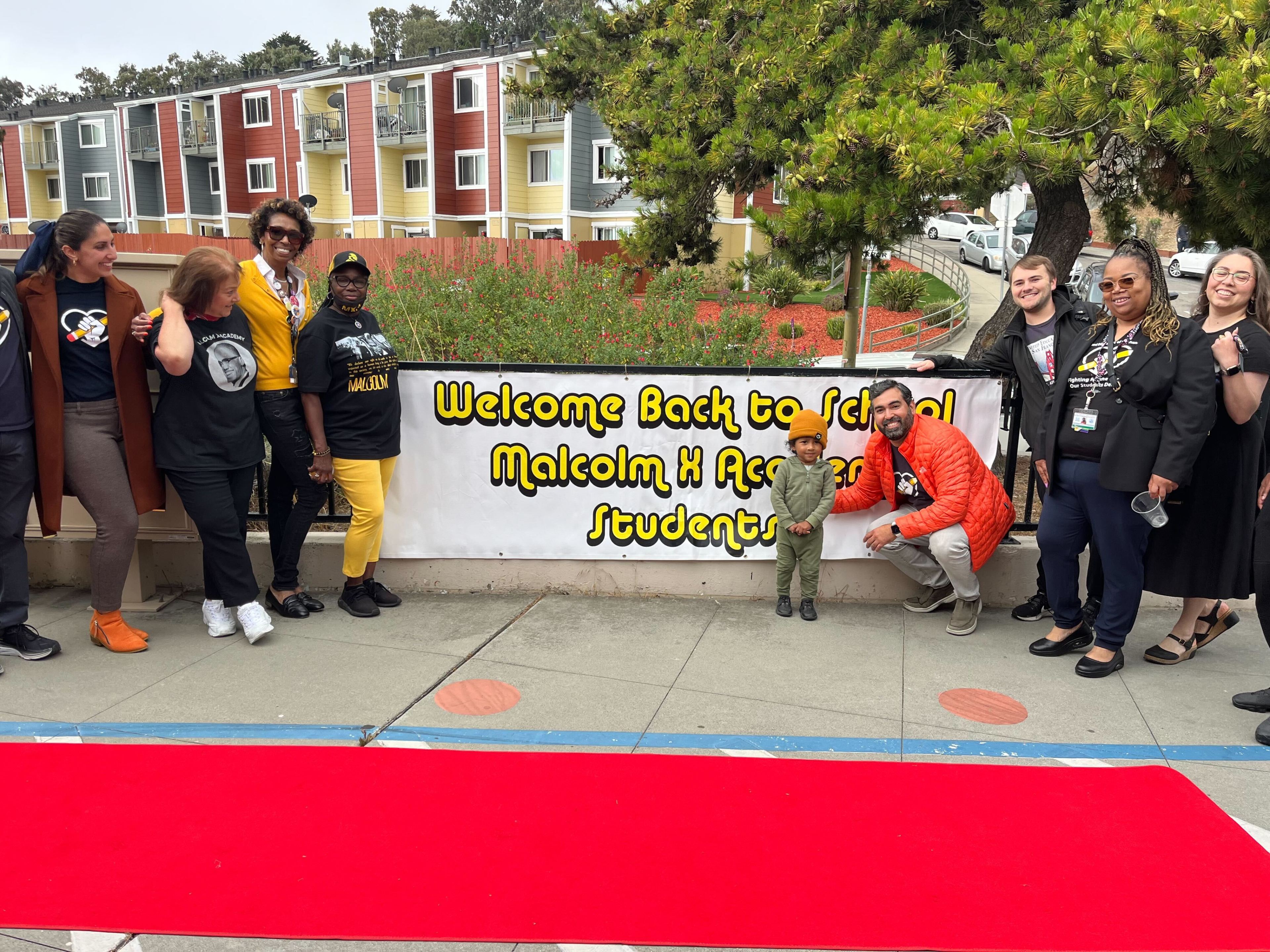 A group of smiling individuals stands near a banner reading &quot;Welcome Back to School Malcolm X Academy Students,&quot; with colorful buildings and greenery in the background.