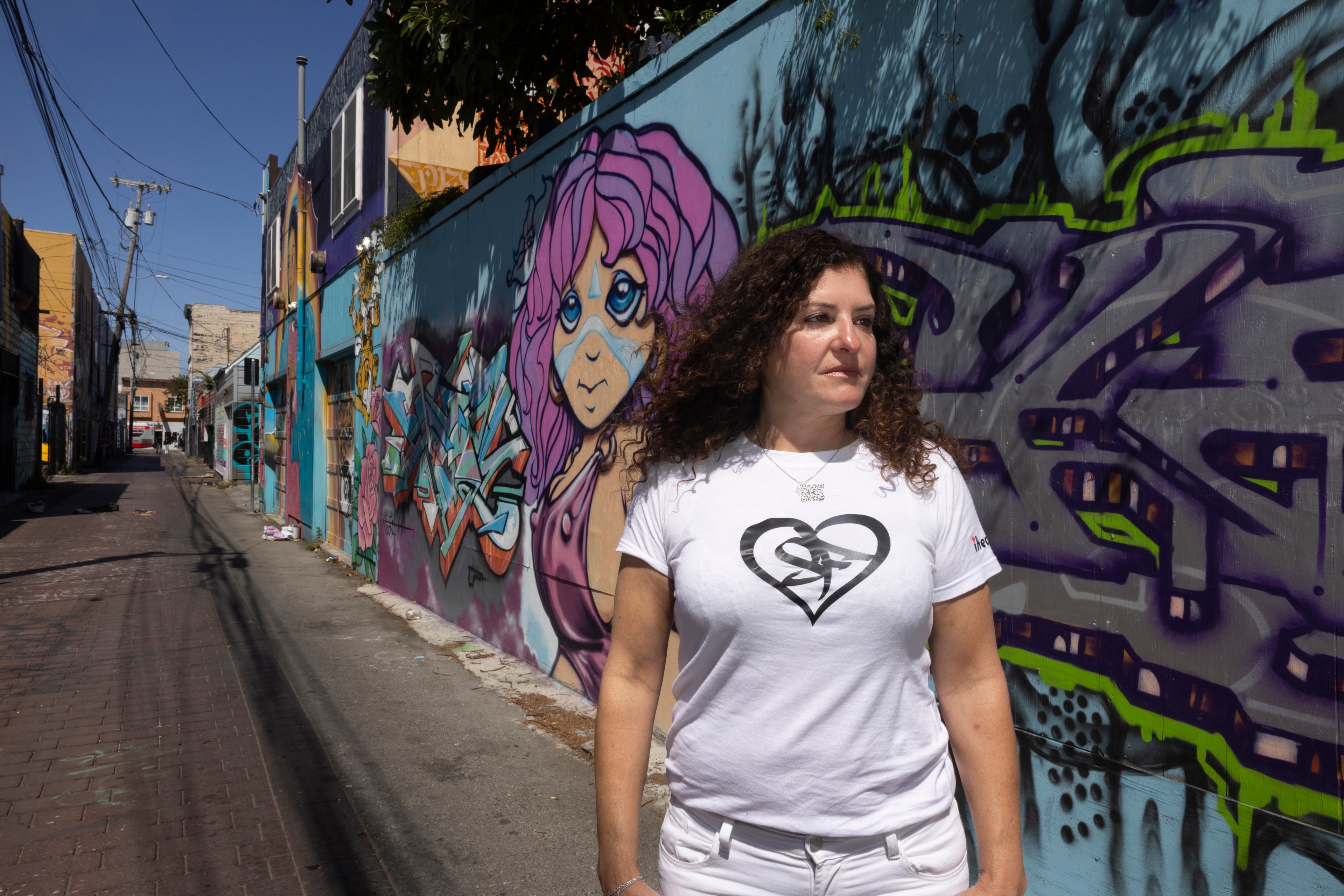 A woman in a white t shirt stands in front of a brightly colored mural