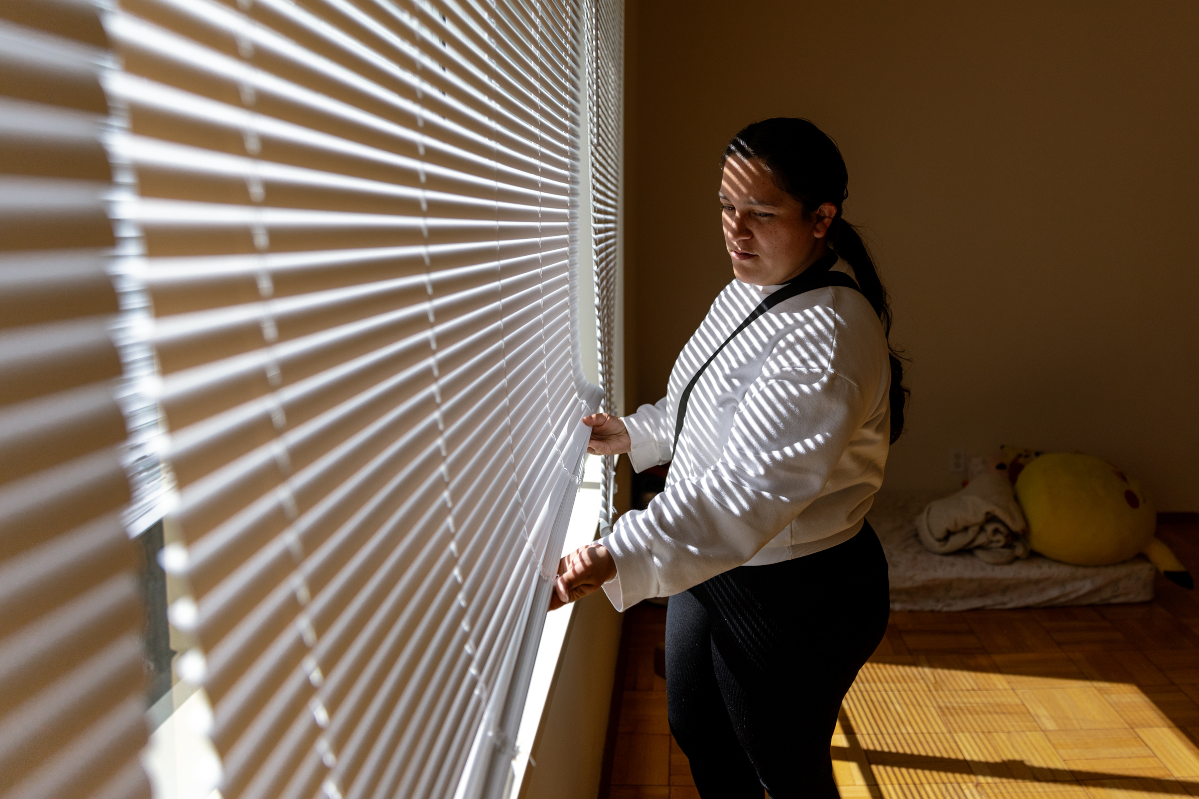 A woman in a white top and black pants stands by a window adjusting the blinds, with sunlight casting striped shadows on her and the room, which has a cushion and yellow pillow on the floor.