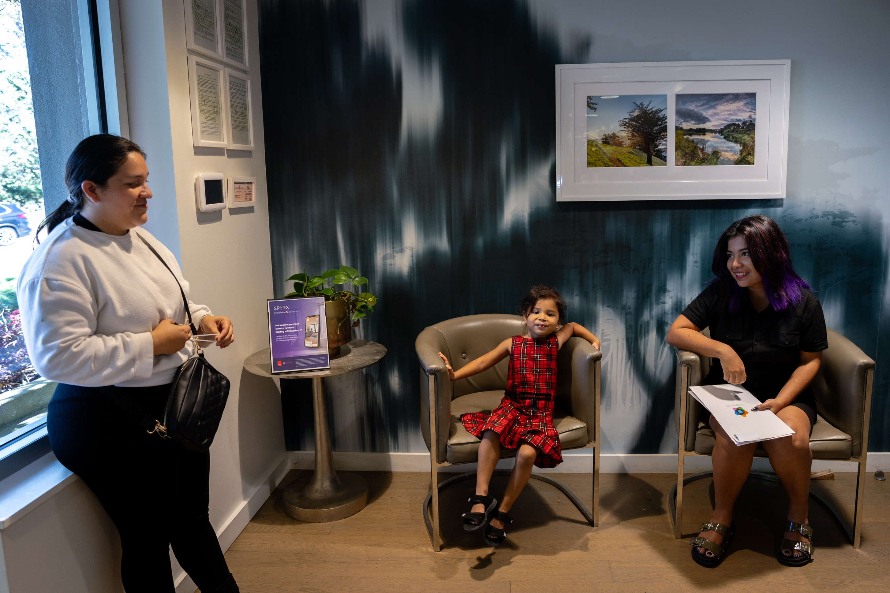 Three people are in a waiting room. A woman stands near a window, while a child in a red dress and a woman with purple hair sit in chairs. A table with a plant and pamphlet is nearby.