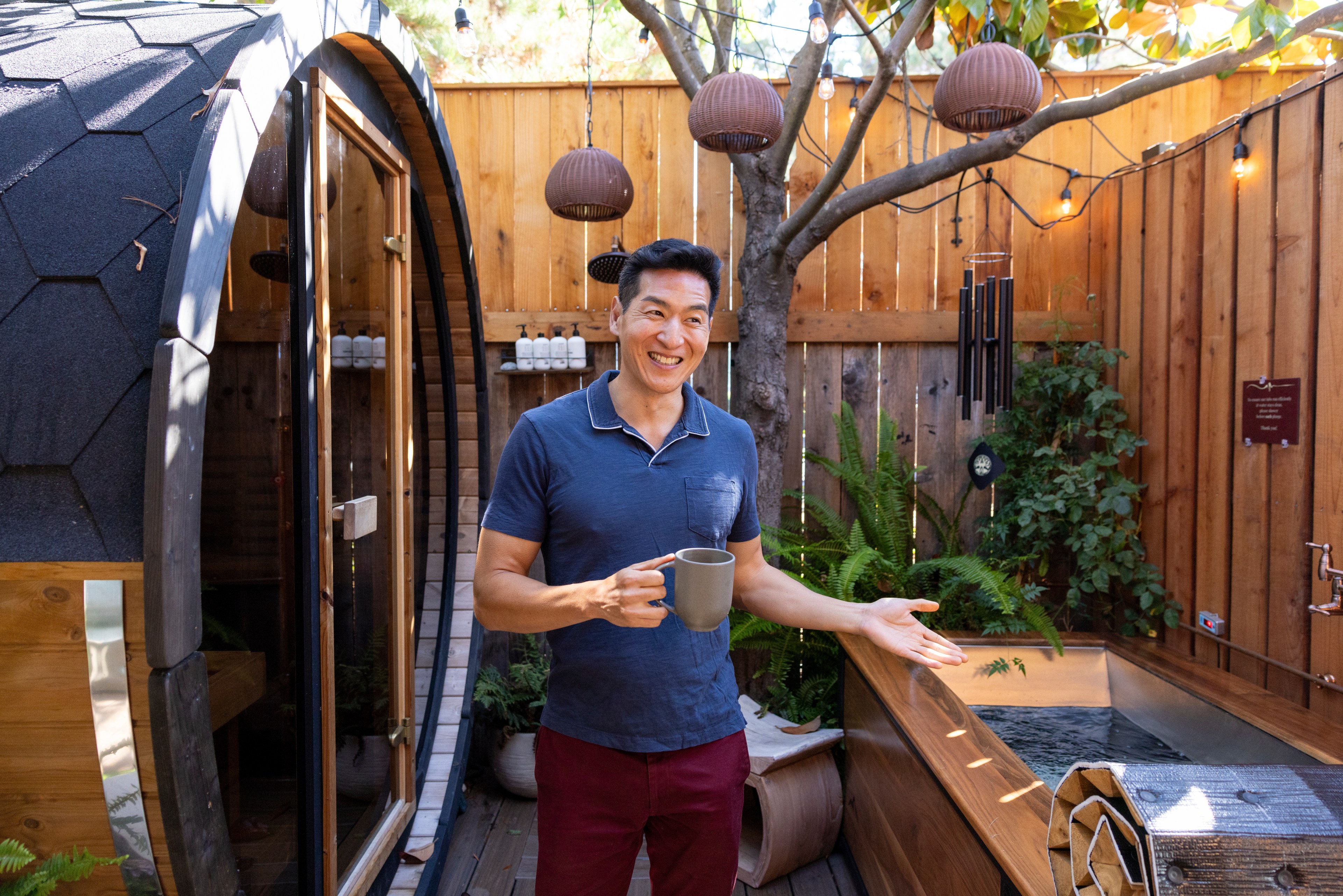 A man with a mug smiles and gestures in a cozy outdoor space with wooden walls, lush plants, a spa tub, and a sauna-like structure behind him decorated with string lights.