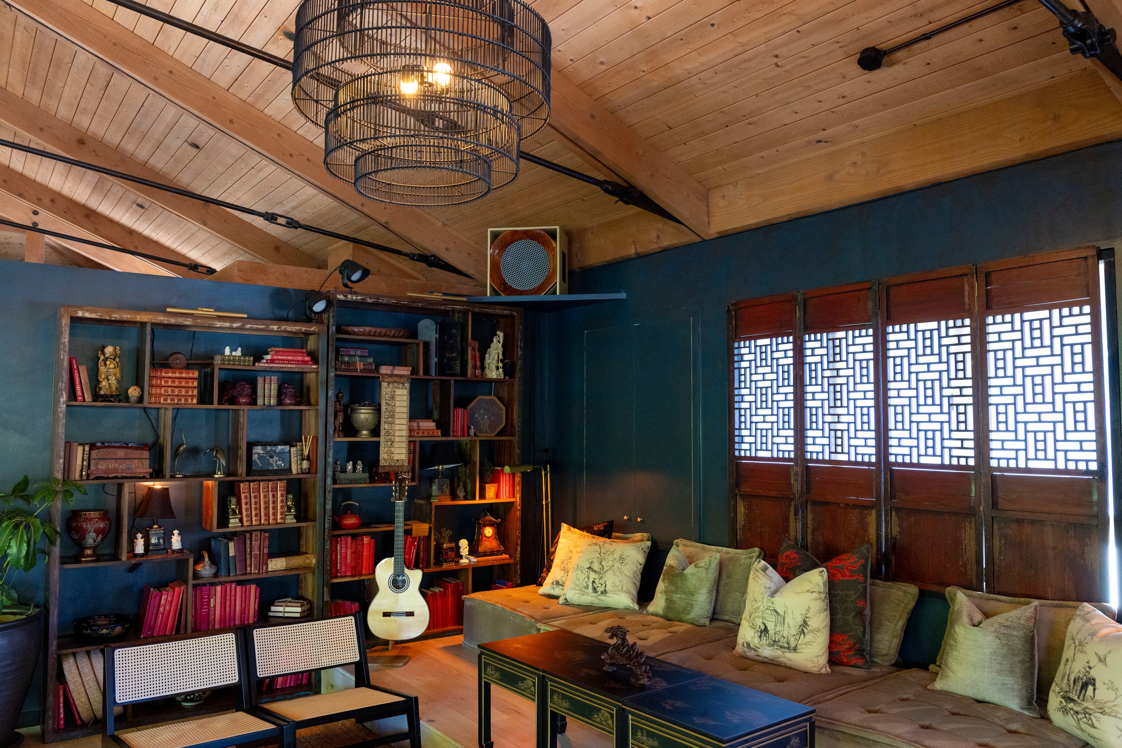 A cozy room with wooden ceilings, bookshelves full of books and decor, a guitar, cushioned bench seating, Chinese lattice windows, and a large metal chandelier.