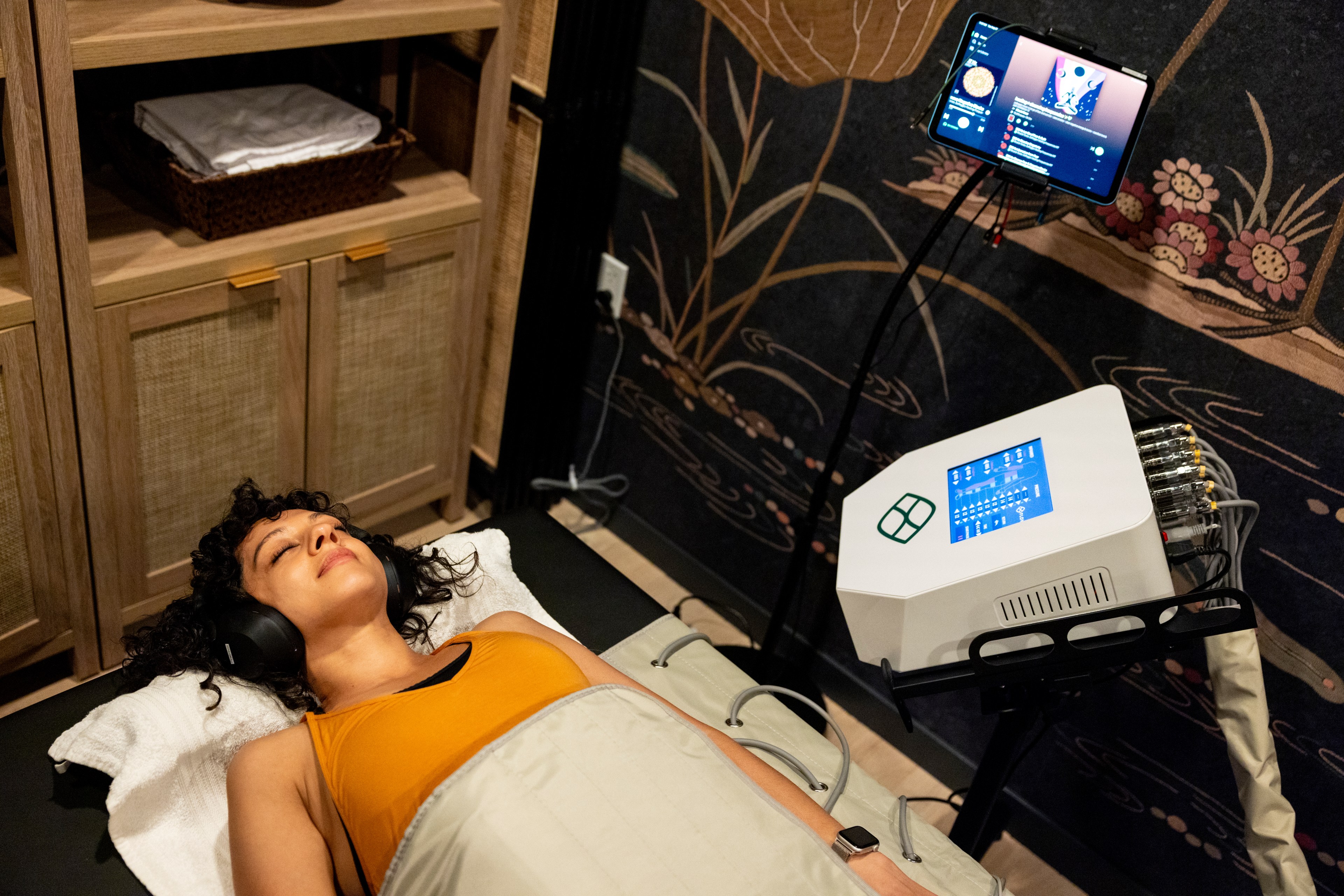 A woman lies on a padded table, wearing headphones and a yellow tank top. She appears relaxed with a machine and screen next to her. The room has wood cabinets and a decorative wall.