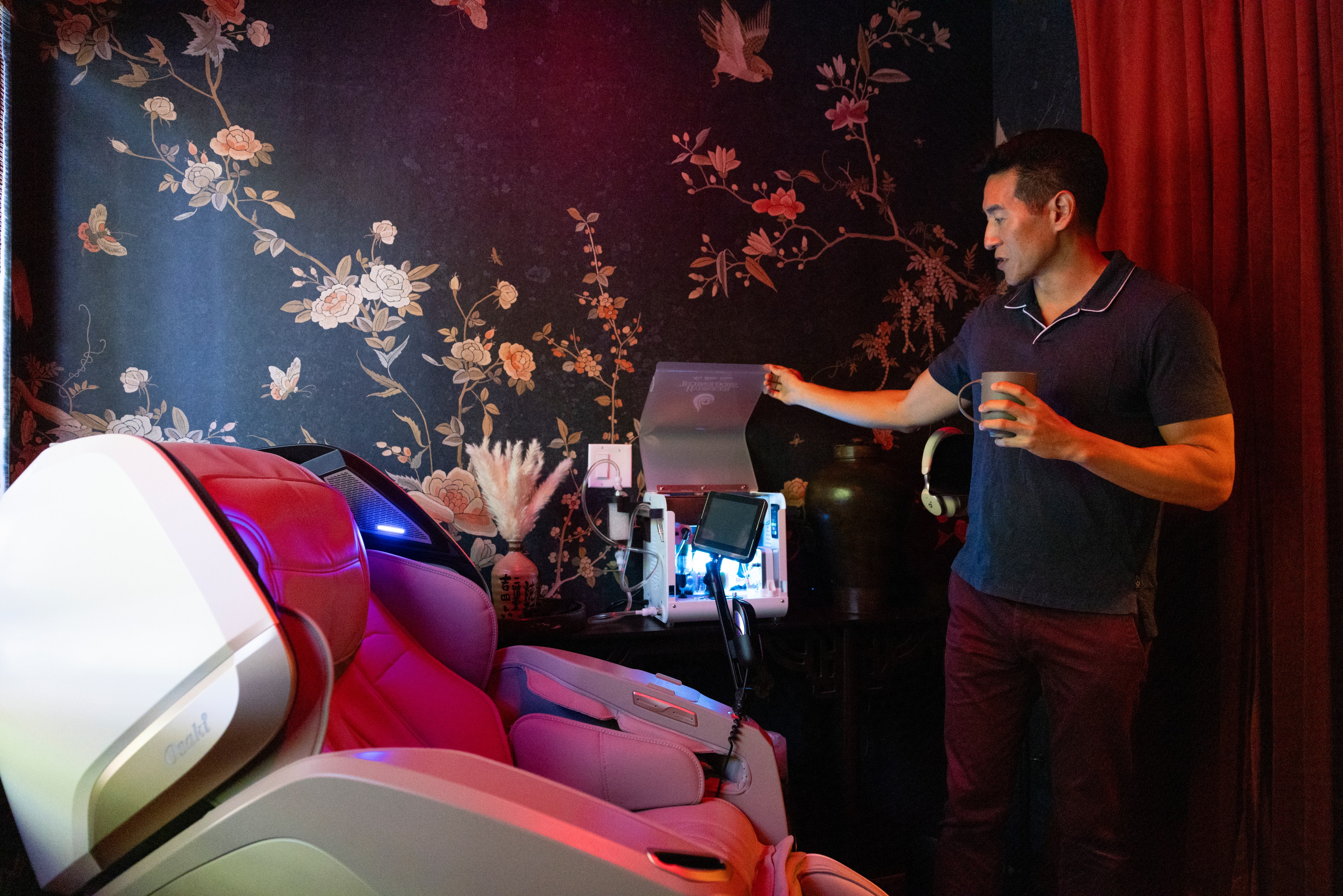 A man stands beside a futuristic, red-lit massage chair, holding a cup, with a floral wallpaper and various gadgets on a table behind him in a dimly lit room.