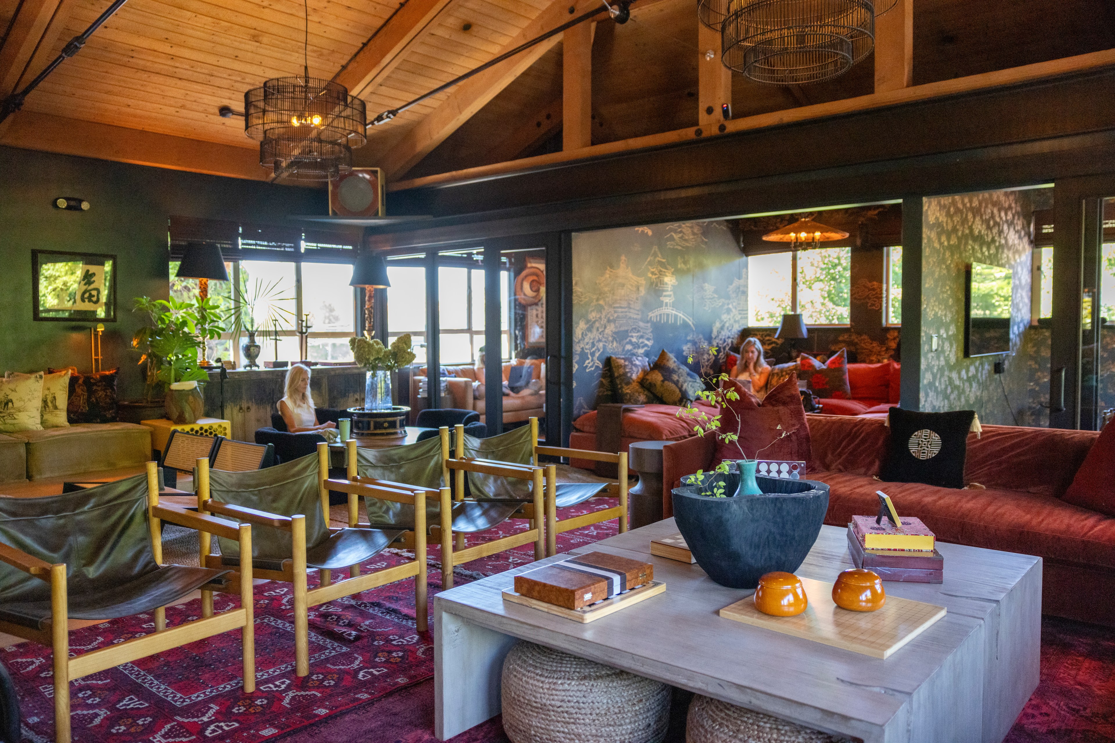 A cozy, rustic living room with wooden ceiling beams, colorful rugs, a mix of red and green seating, plants, and large windows displaying natural light.