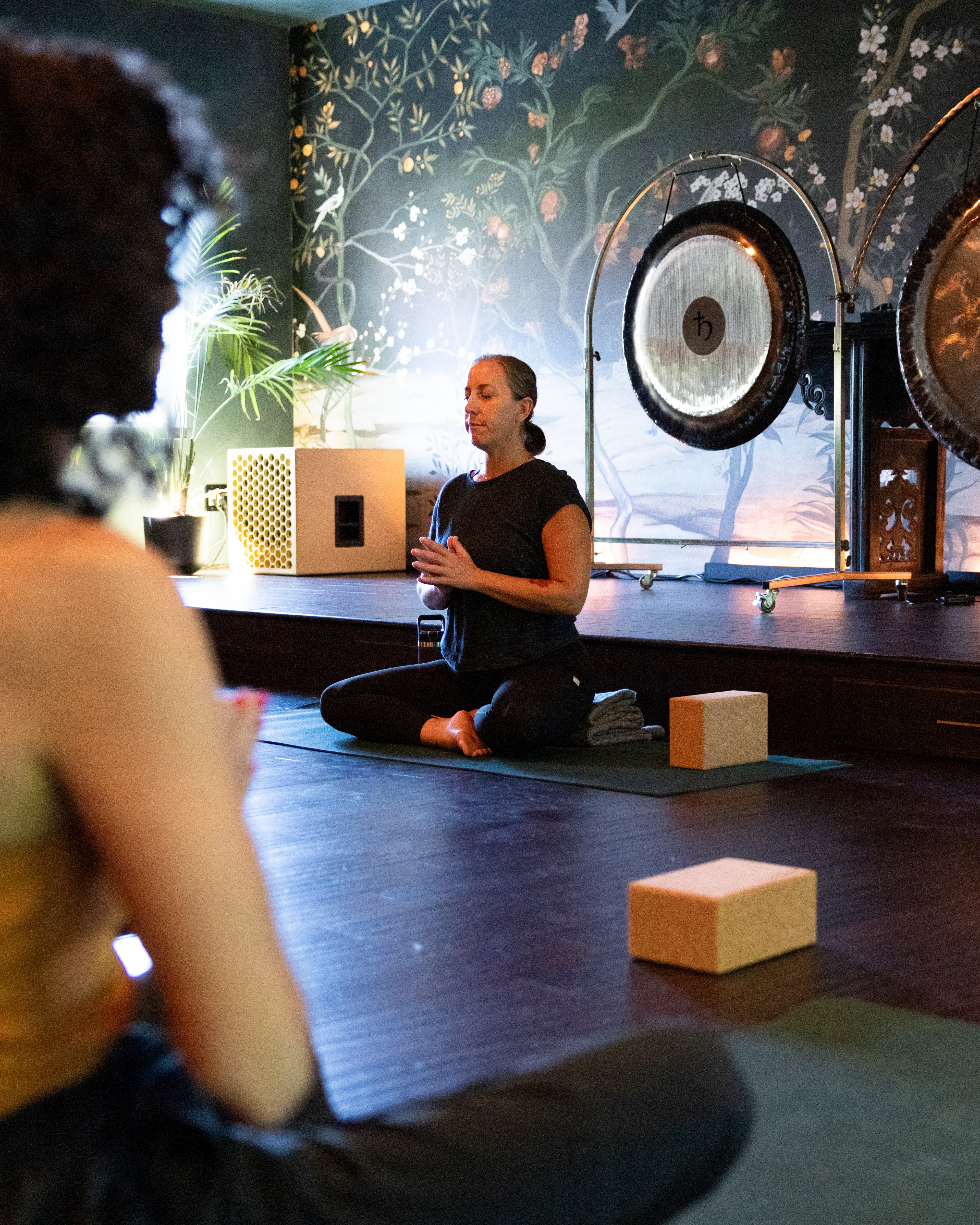 A woman sits cross-legged in a meditation pose on a yoga mat, with another person in the foreground. The room has floral wallpaper and large gongs.