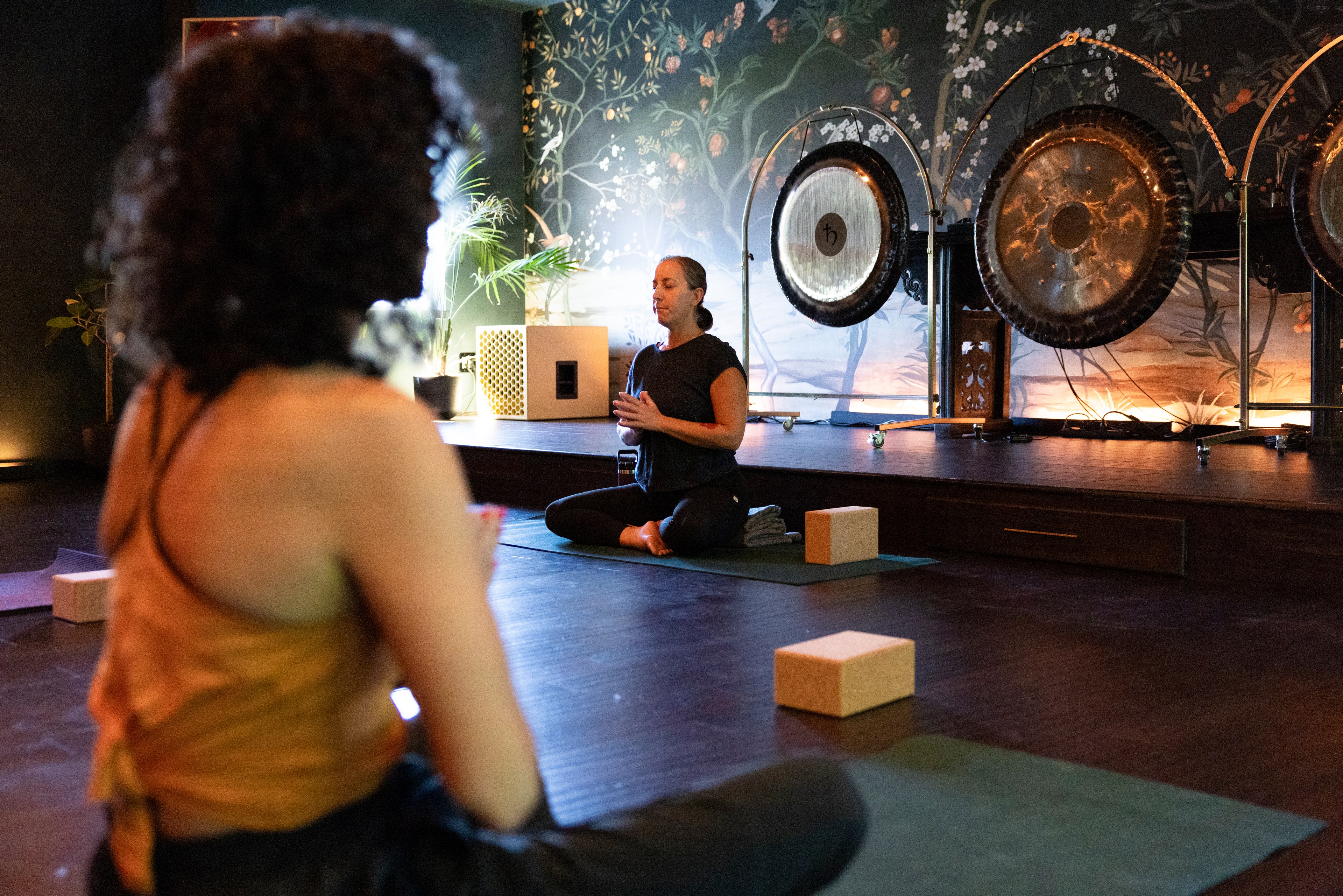 Two people sit on yoga mats in a dimly lit room. One faces the camera in the foreground, while the other meditates near large gongs and a mural in the background.