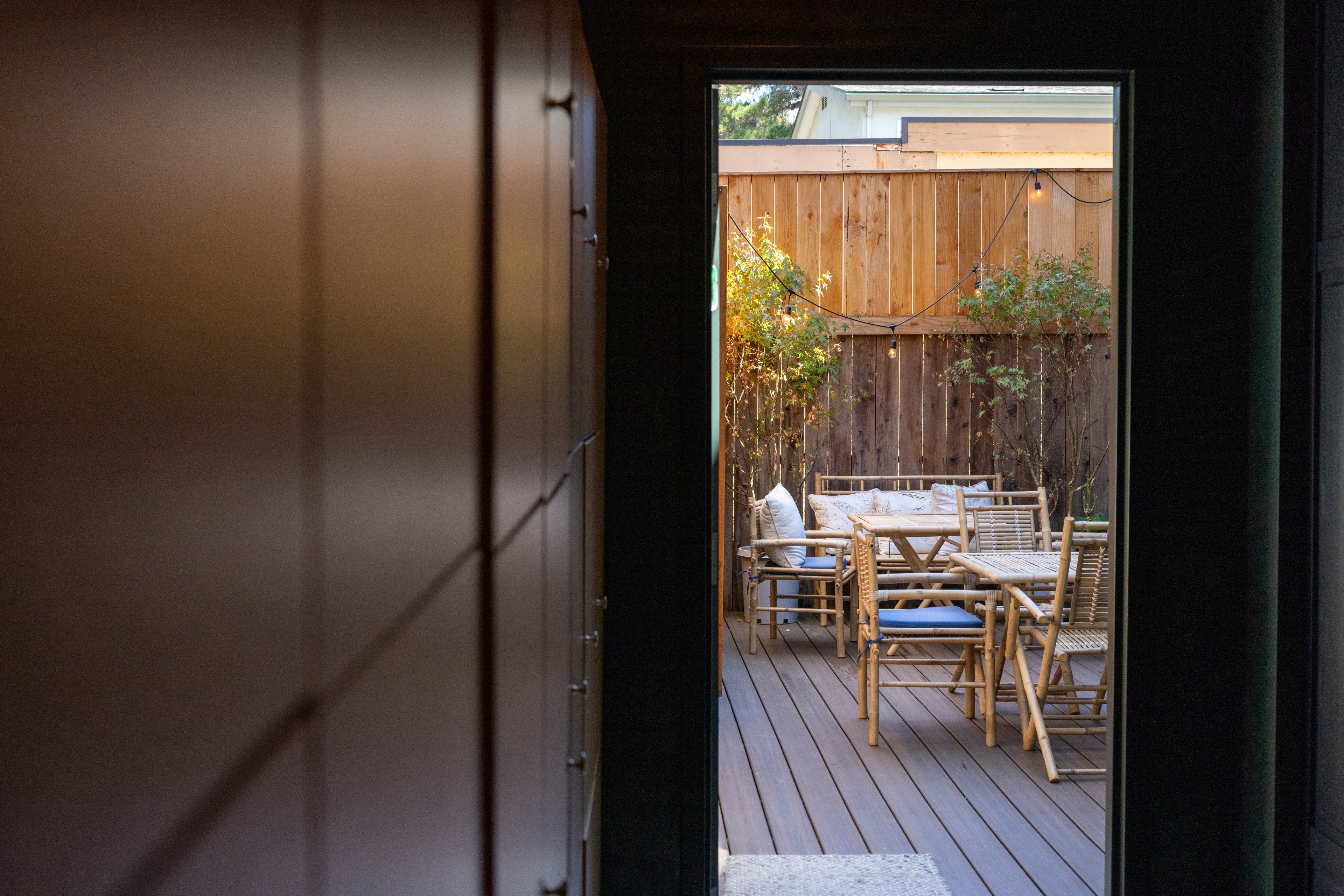 The image shows a cozy outdoor area through a doorway, featuring wooden furniture, potted plants, string lights, and a wooden fence, creating a warm and inviting space.