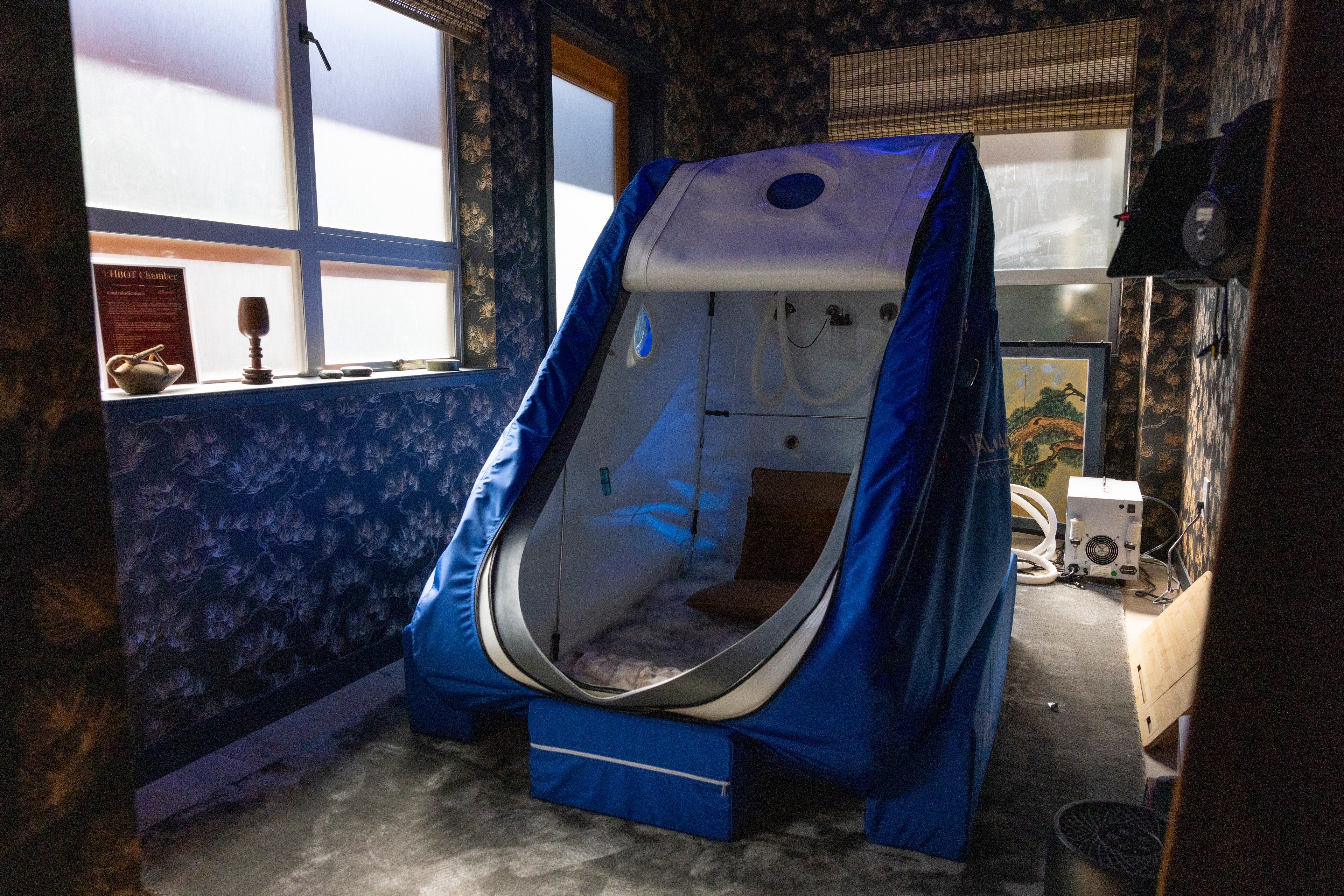 The room contains a blue hyperbaric oxygen chamber next to a window with floral wallpaper and various small objects on the sill. The chamber is partially open, revealing its interior.