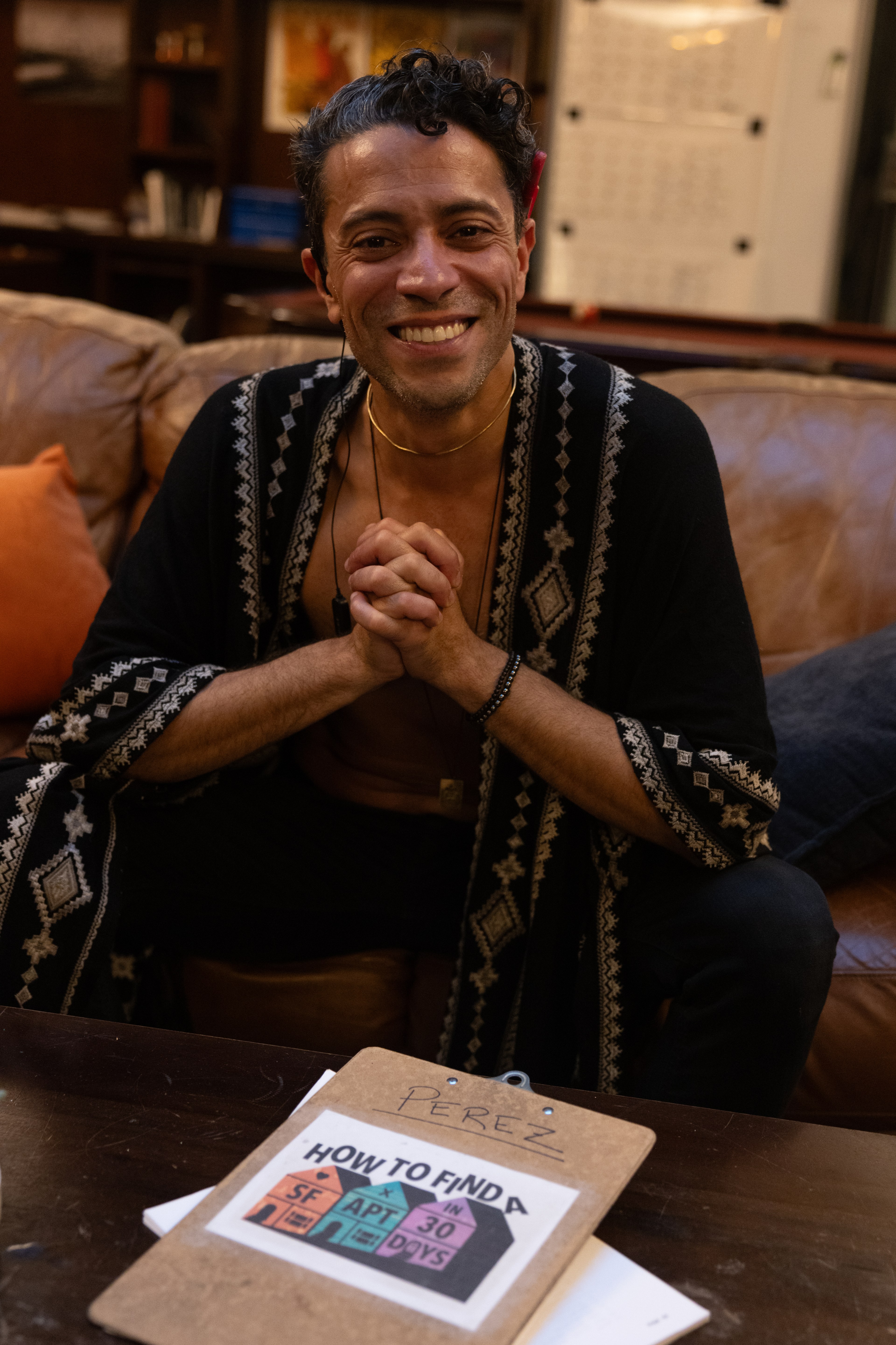 A smiling person sits on a couch with hands clasped. In front, there's a clipboard labeled &quot;PEREZ&quot; with a guide titled &quot;How to Find a SF Apt in 30 Days.&quot;