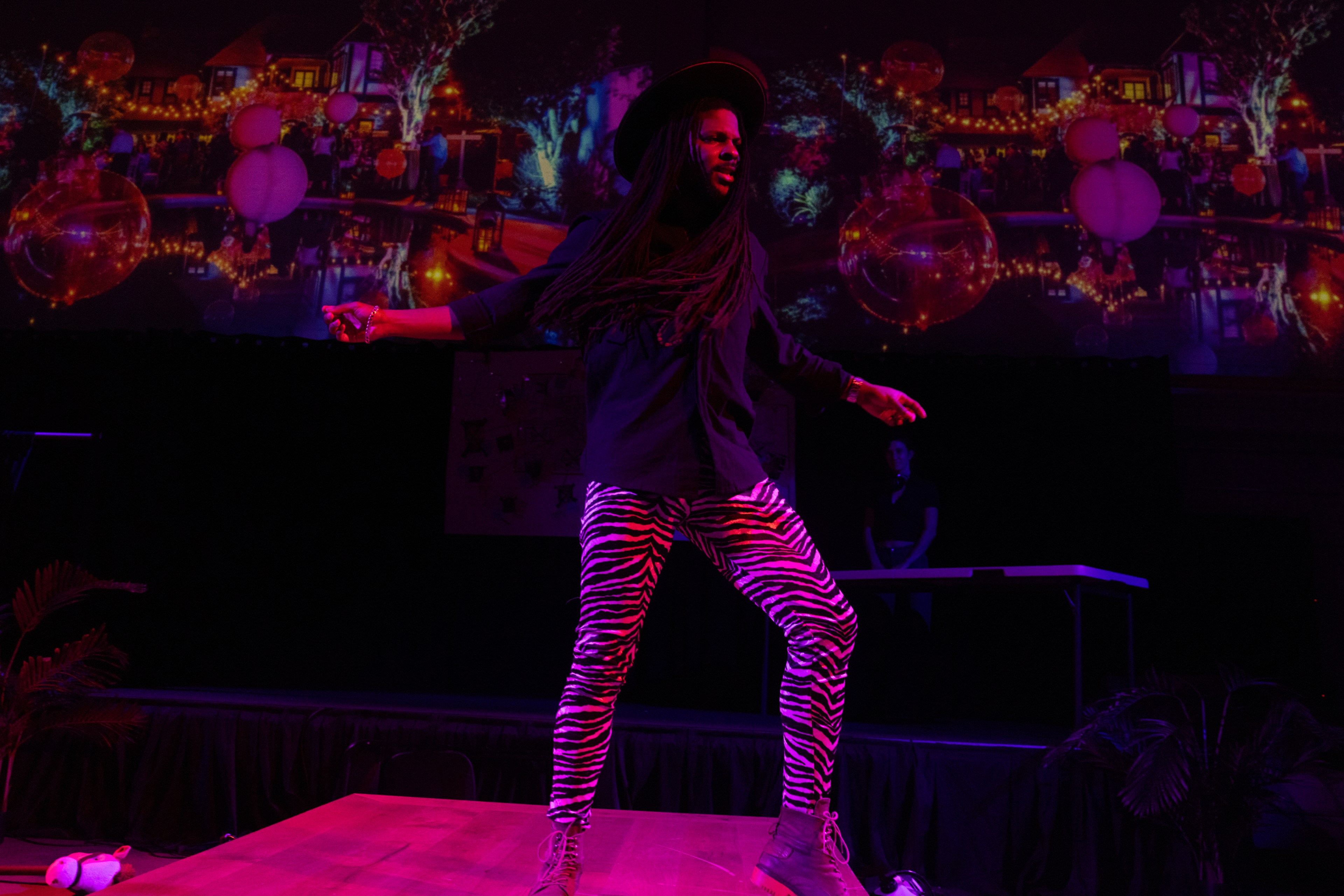 A person with long hair wearing a black hat, black shirt, and zebra-print pants is performing on a stage with a colorful, festive background projection.