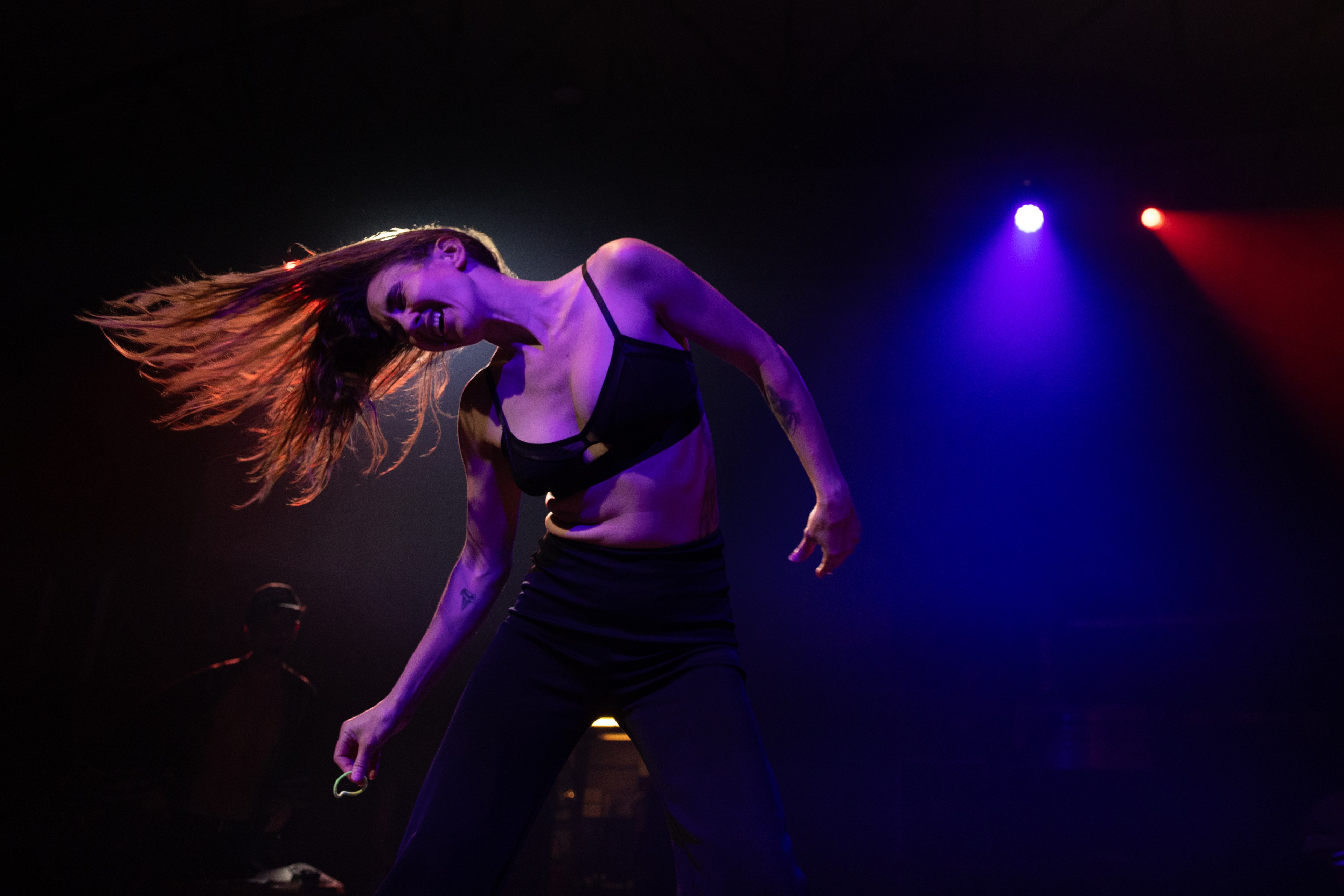 A woman is dancing energetically on a stage under colorful lights; her head is tilted, and her hair is flipping to the side. She is wearing a black outfit.