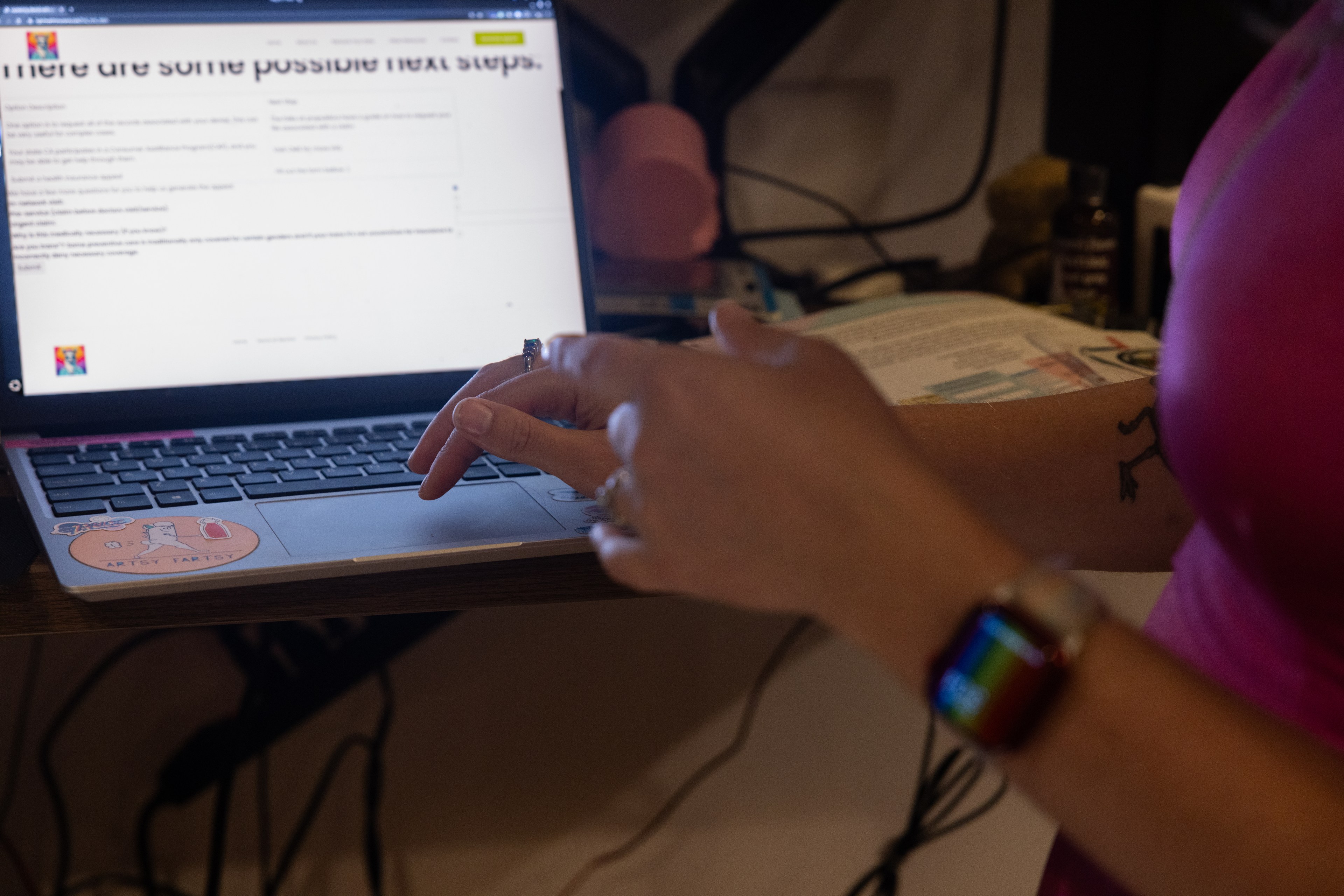 A person wearing a pink shirt and a smartwatch is working on a laptop. They are using their right hand to point at the screen and their left hand is resting on the laptop.