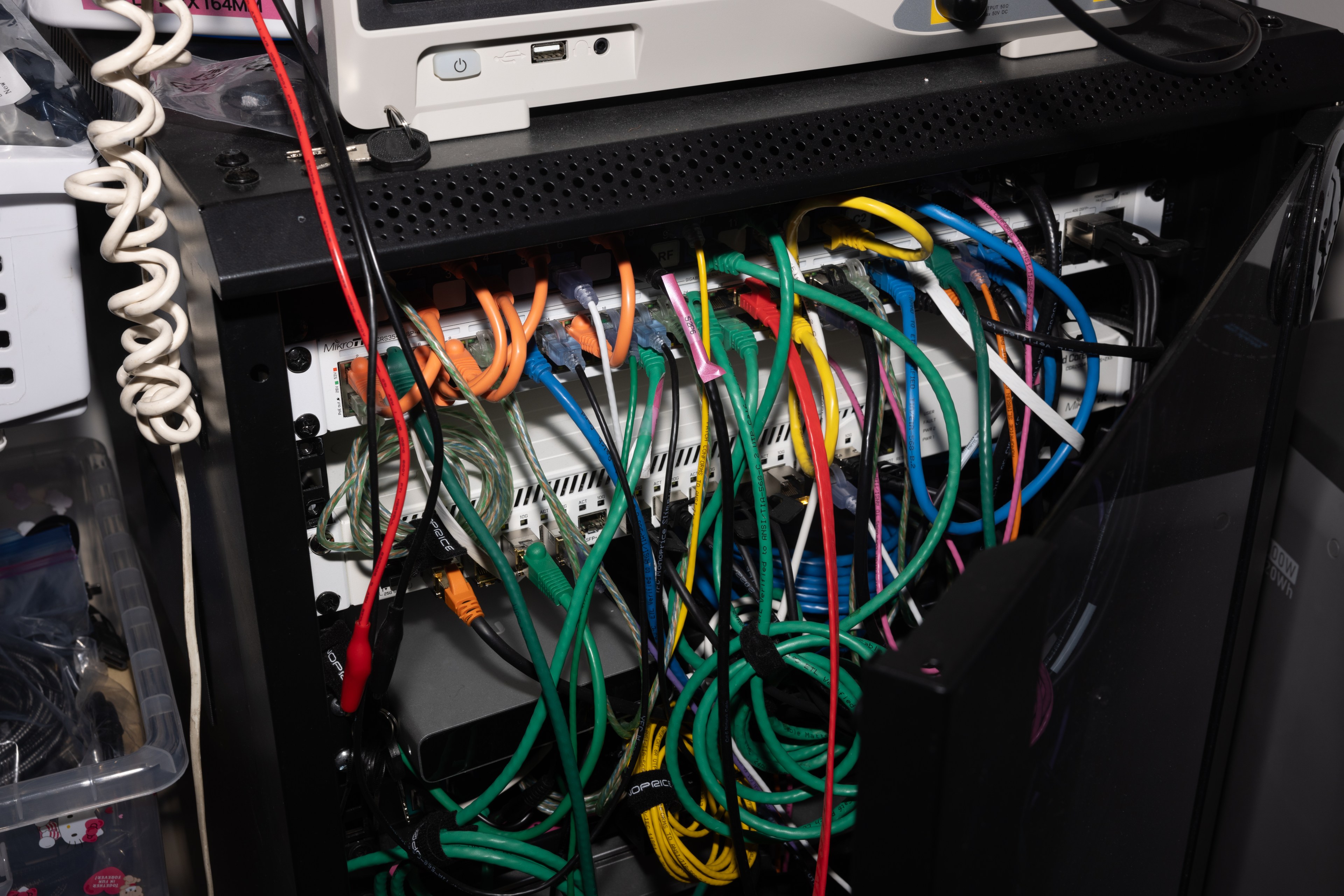 The image shows a tangle of multicolored cables connected to networking devices in a server rack, with wires coiled around various points and additional equipment nearby.