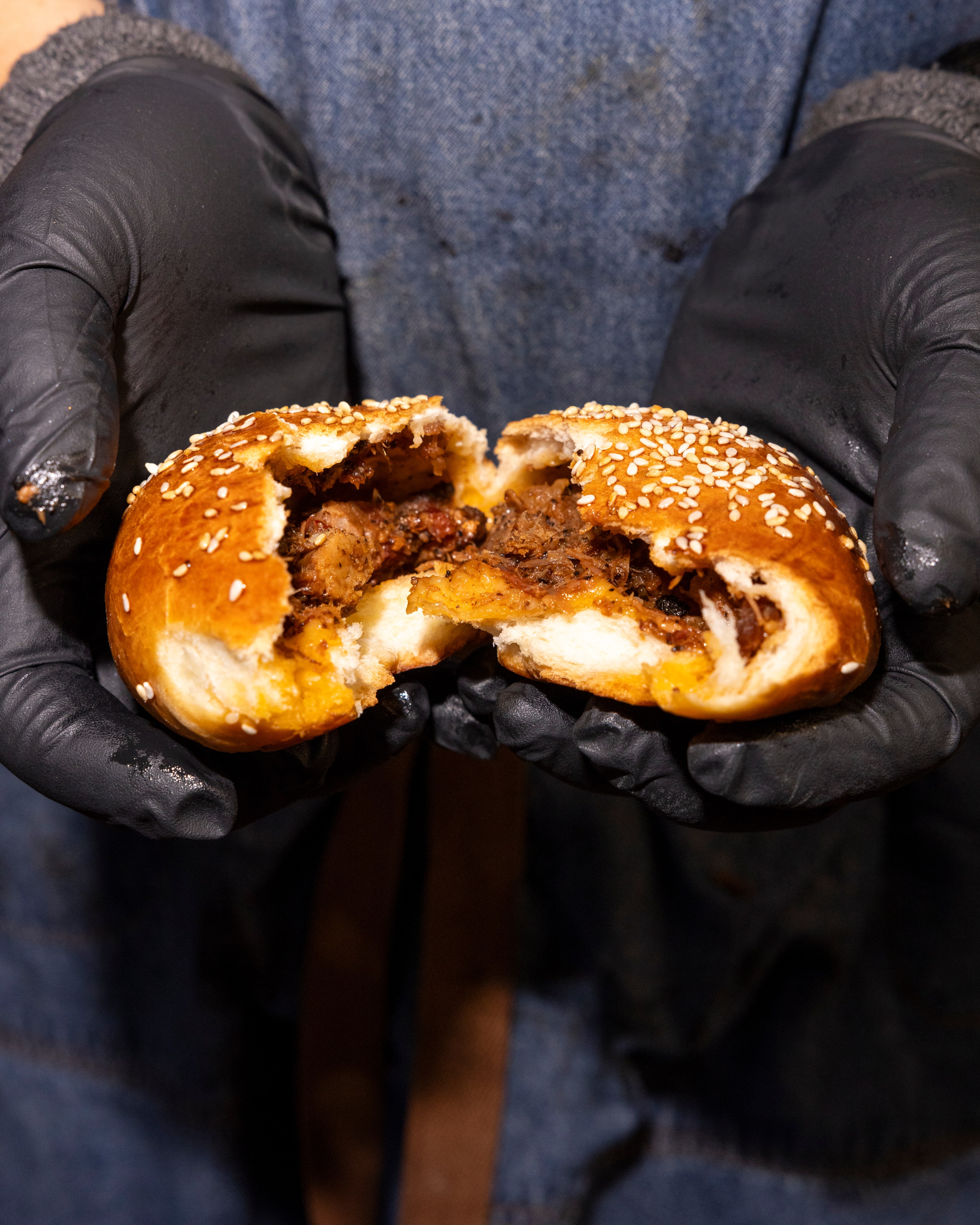 A person wearing black gloves is holding a sesame seed bun filled with pulled meat and melted cheese, torn open to show the tasty contents.