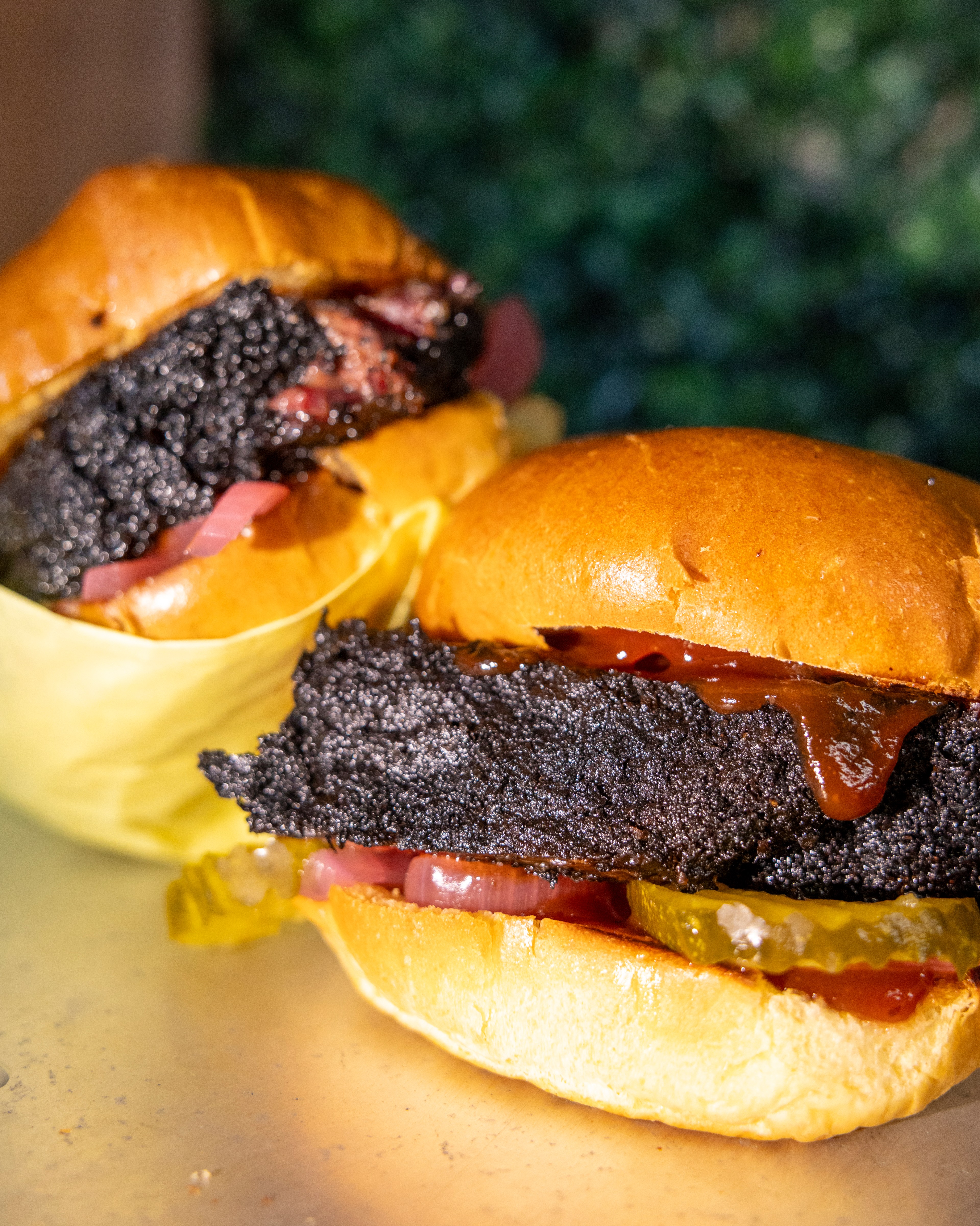 The image features two burgers with crispy, charred meat, pickles, onions, and sauce on shiny, golden buns on a light surface with a greenery background.