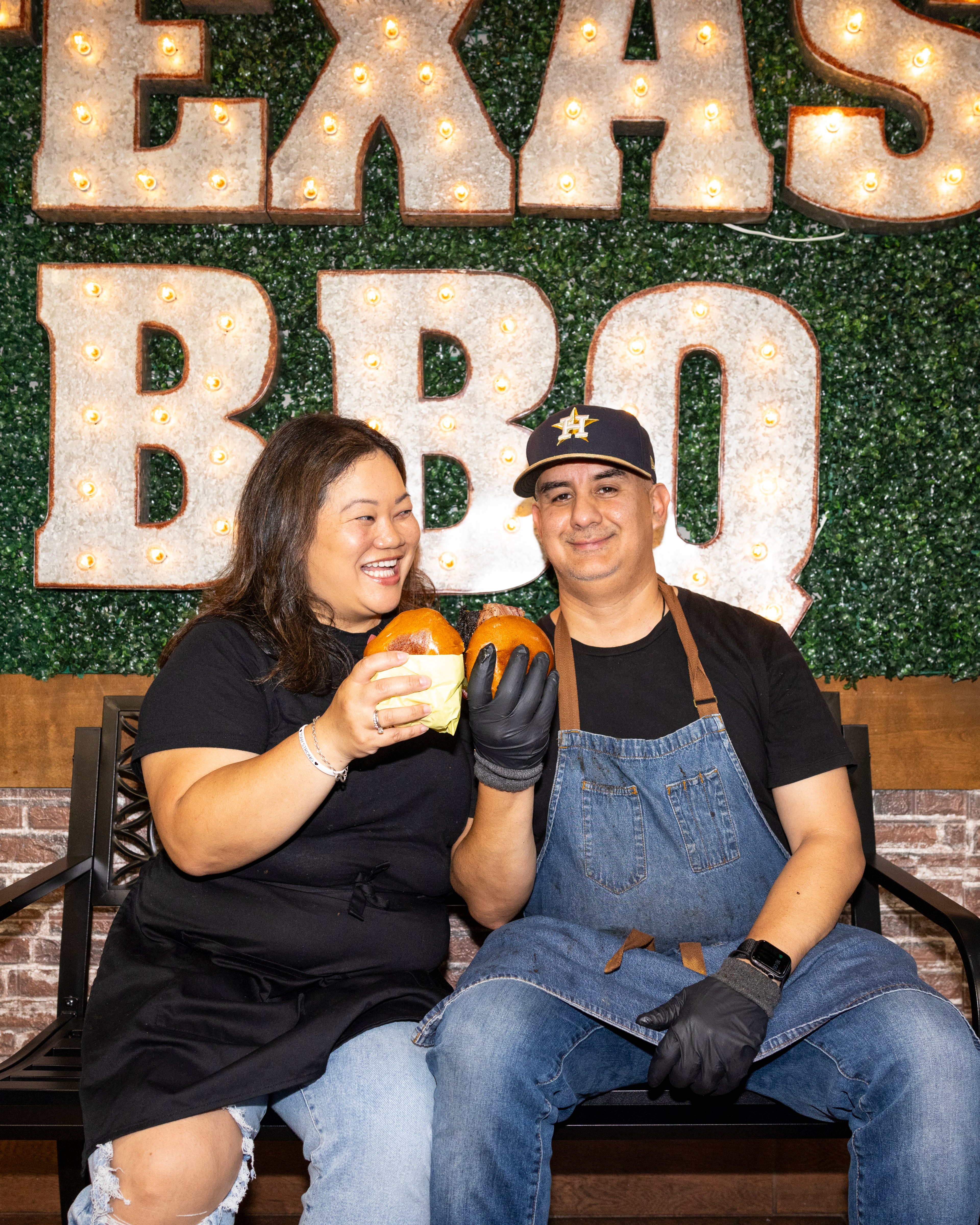 A smiling woman and a man in an apron hold burgers in front of a "TEXAS BBQ" sign with lights. They are sitting on a bench with a green background and brick detail.