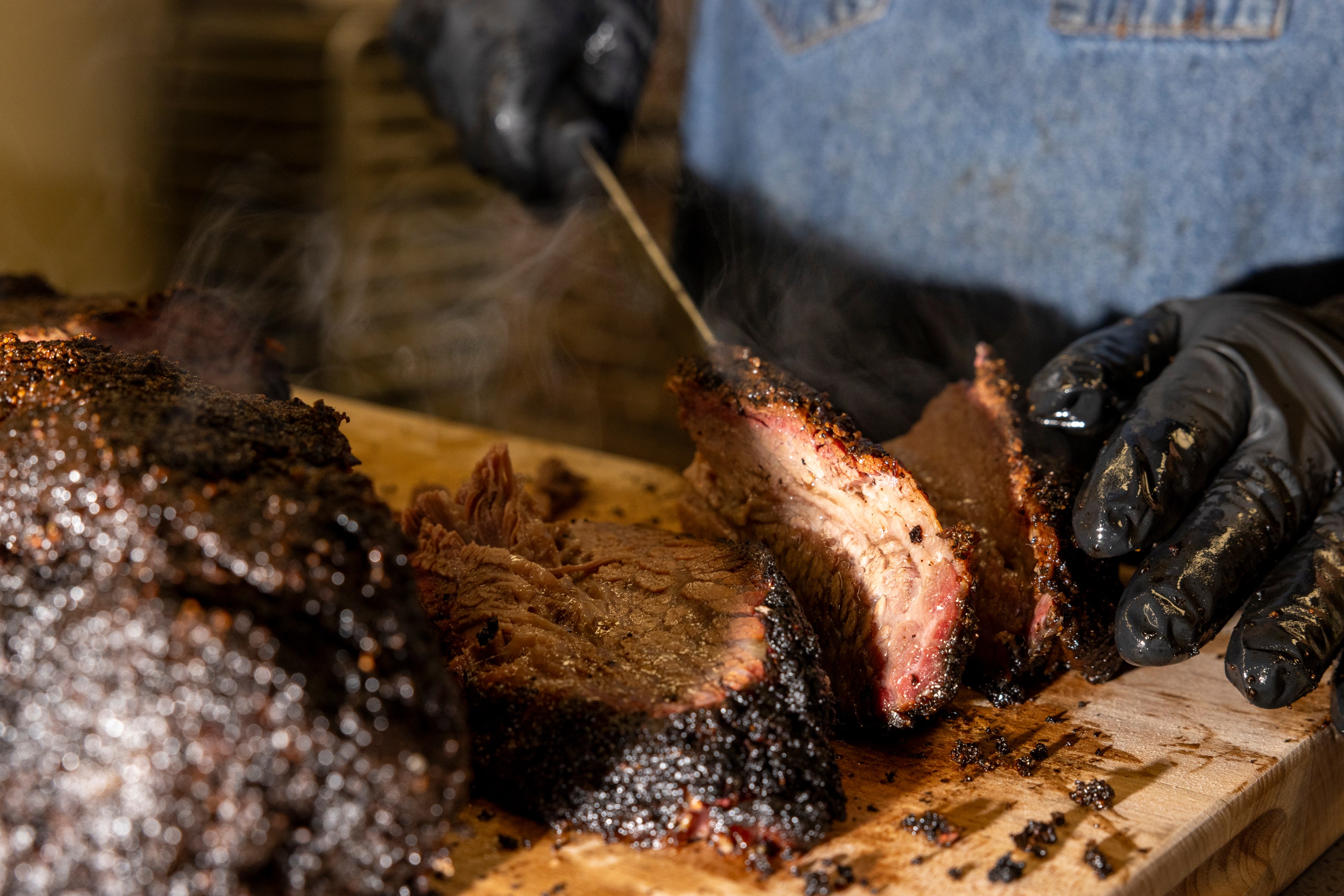 A person wearing black gloves is slicing a piece of steaming, well-cooked brisket on a wooden cutting board, with the meat appearing juicy and tender.