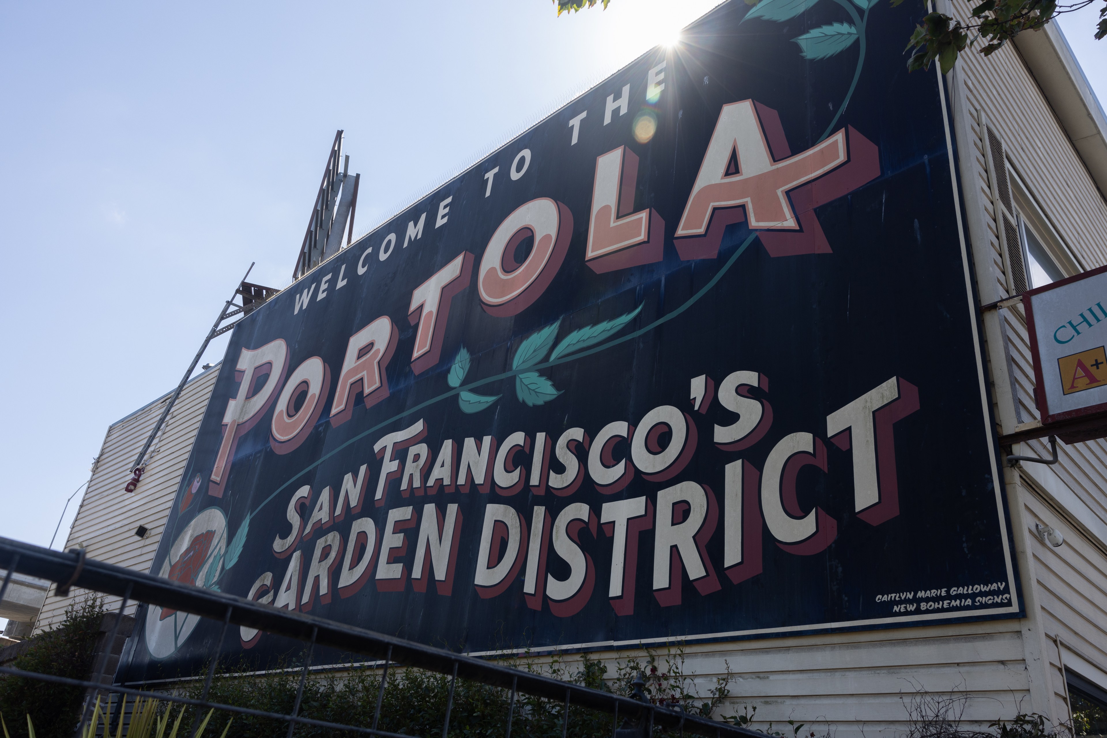 A large sign reads, &quot;Welcome to the Portola, San Francisco's Garden District.&quot; It's mounted on a building with the sun shining above.