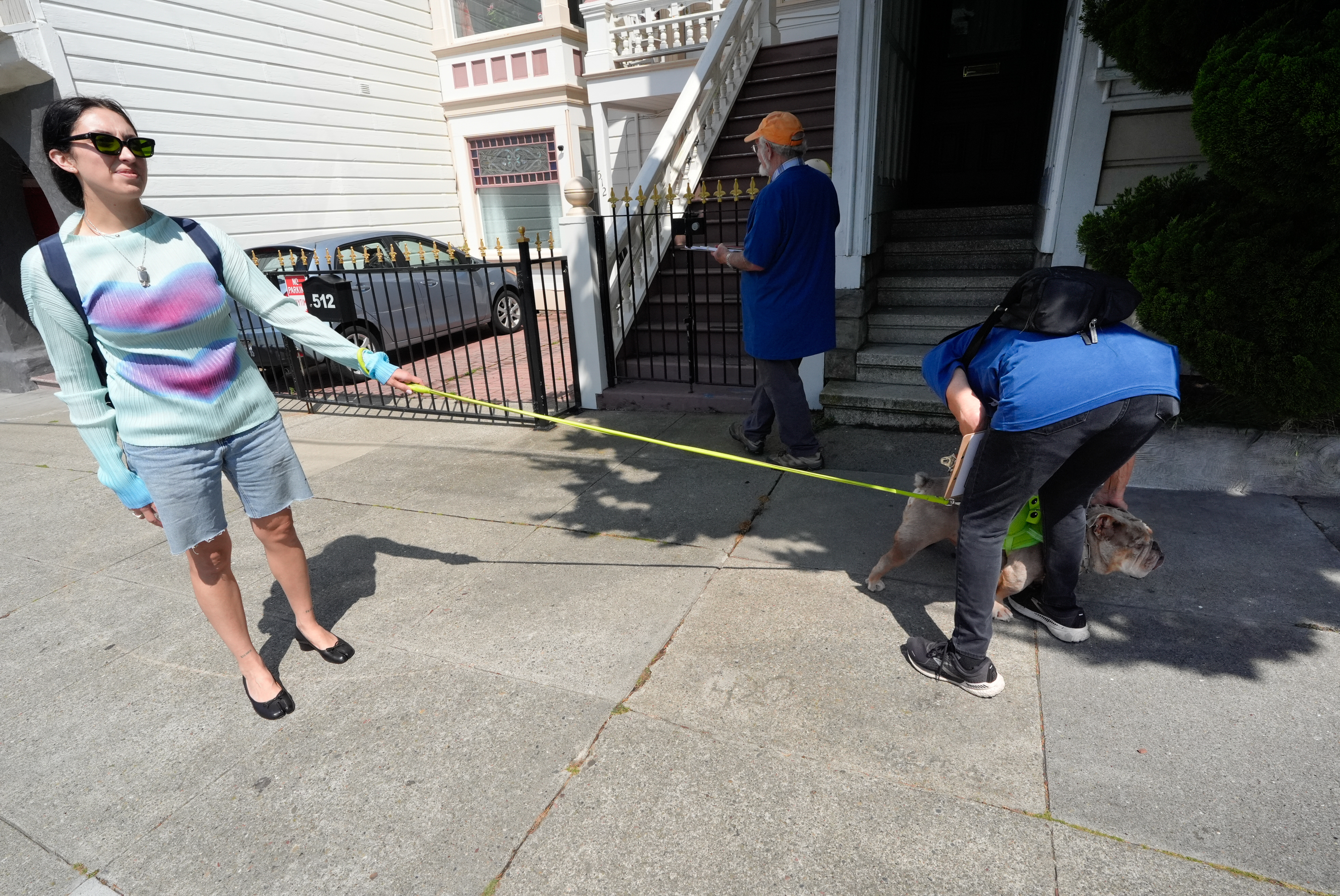 A woman holds a dog leash while standing on a sidewalk. Another person bends down to assist the dog. A third person looks on. They are in front of a gate and stairs.