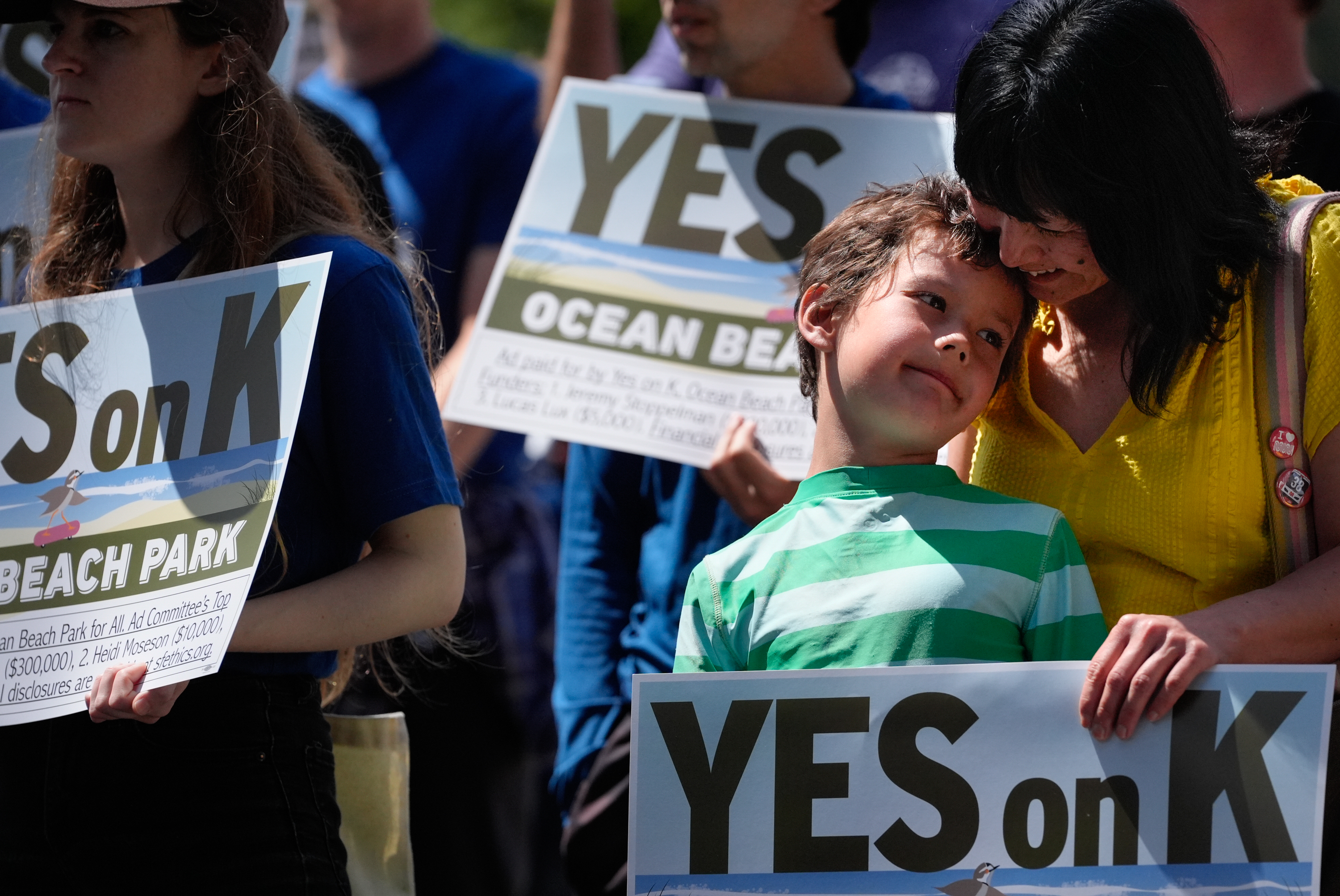 People at a rally hold signs reading &quot;YES on K&quot; with &quot;OCEAN BEACH PARK&quot; in the background. A woman in yellow affectionately leans on a boy in a green-striped shirt.