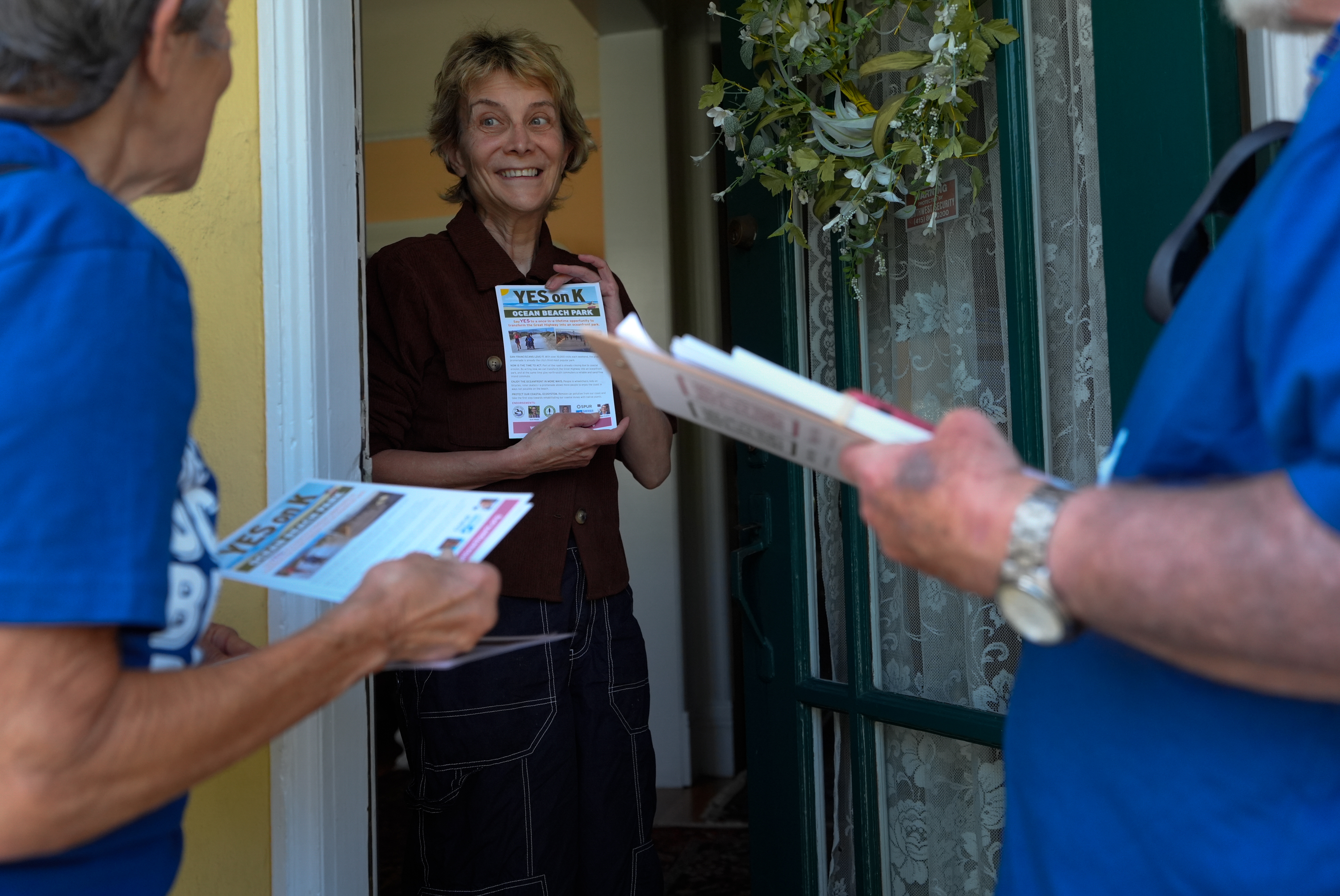 Two people hand out flyers to a third person at her door. She smiles while holding a flyer. The door has a lace curtain and floral wreath hanging.