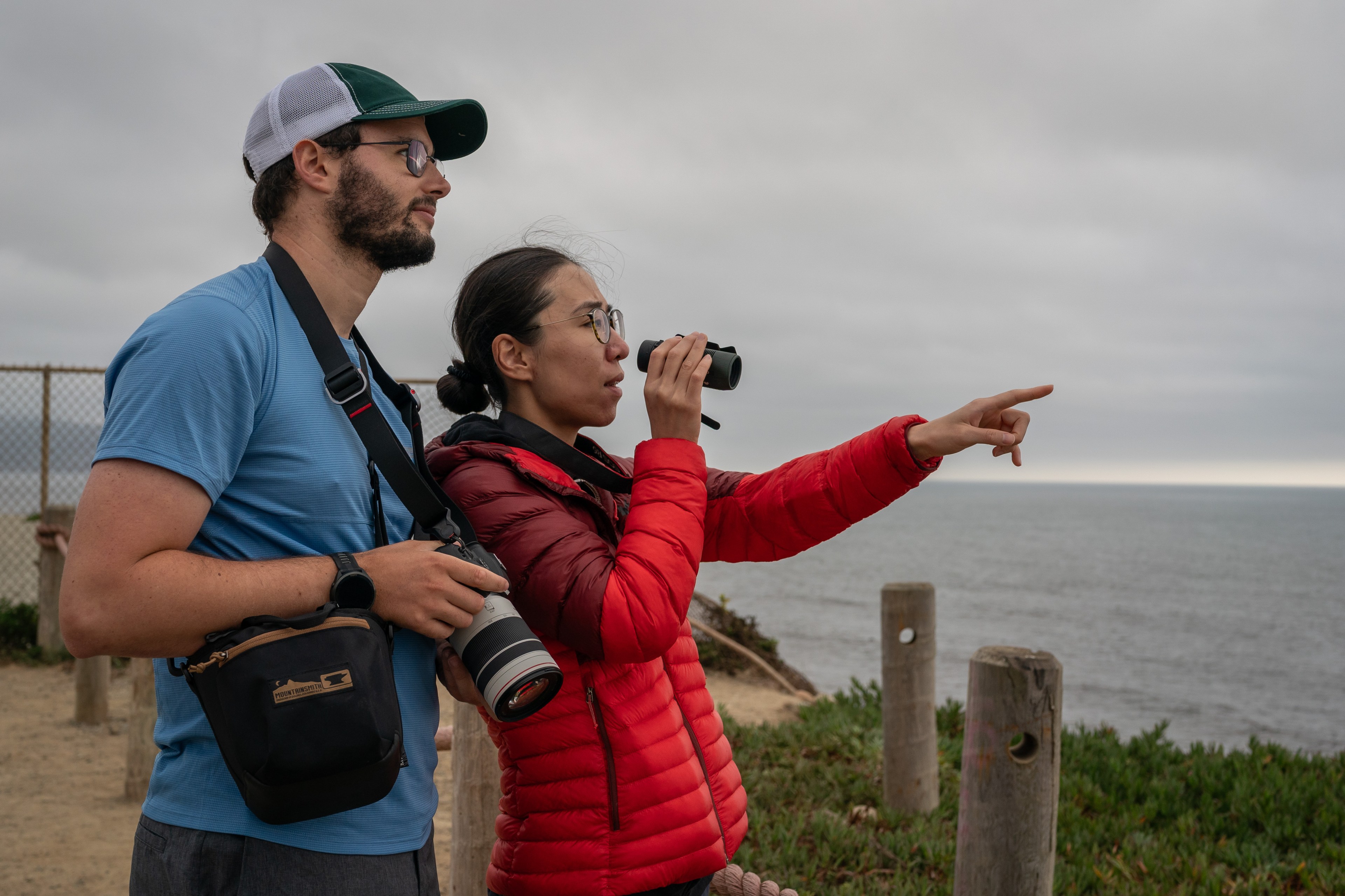 Two people with cameras look at the ocean; one holds binoculars and points while the other carries a camera with a long lens. They're on a cliffside path.