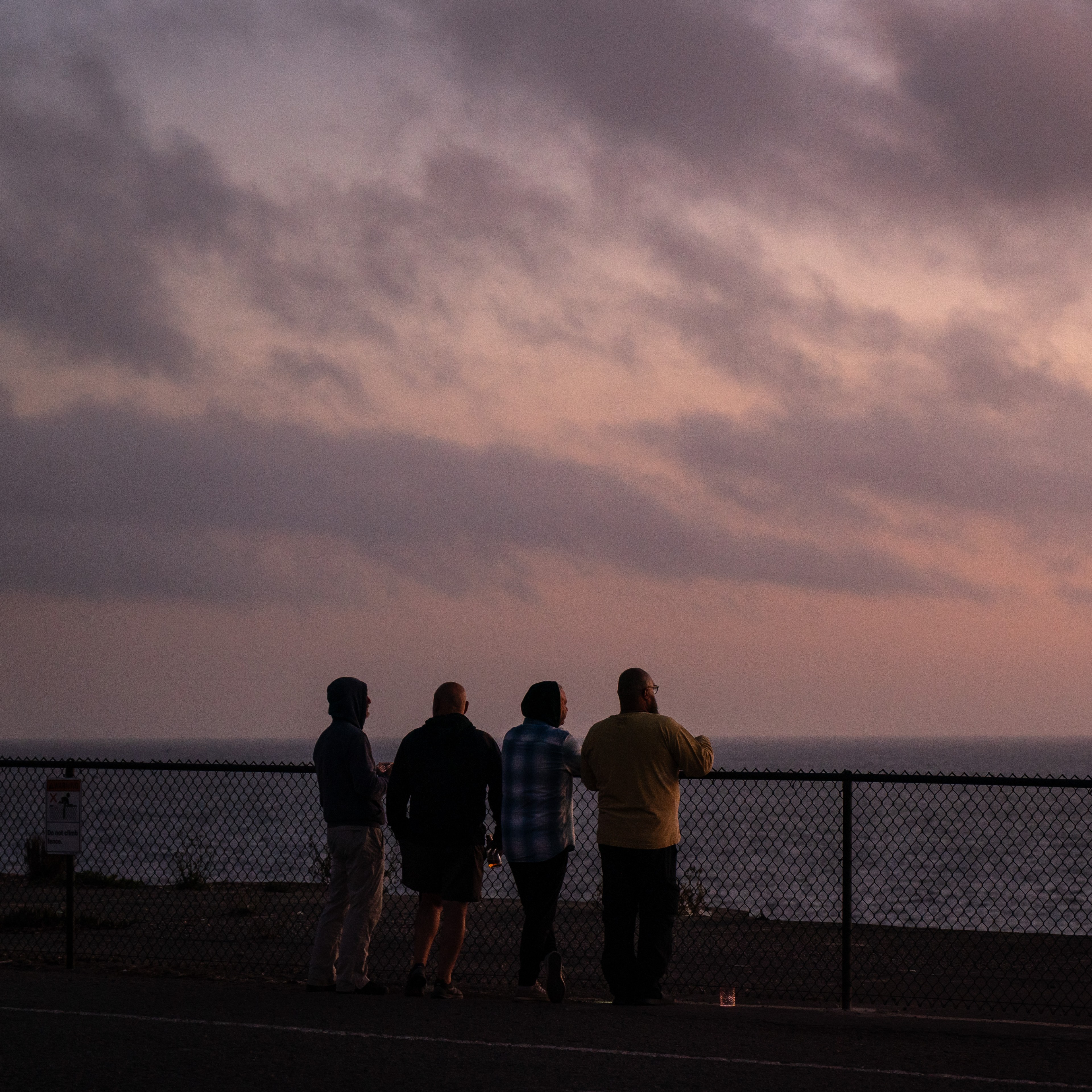 Four people stand beside a fence, gazing at an ocean during sunset under a cloudy sky, their silhouettes highlighted against the dusky backdrop.